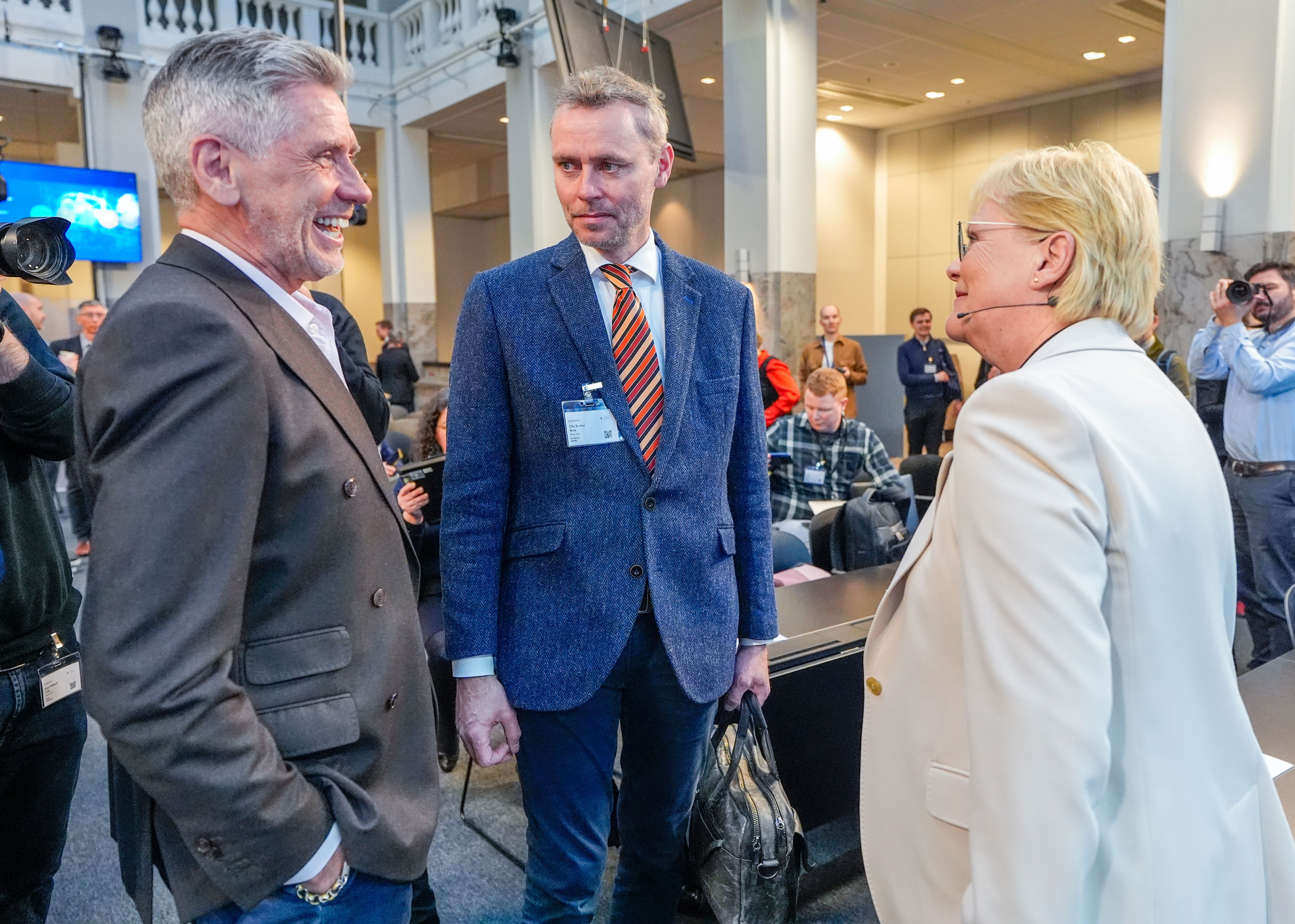 Two men and a woman smiling and talking at an indoor event.