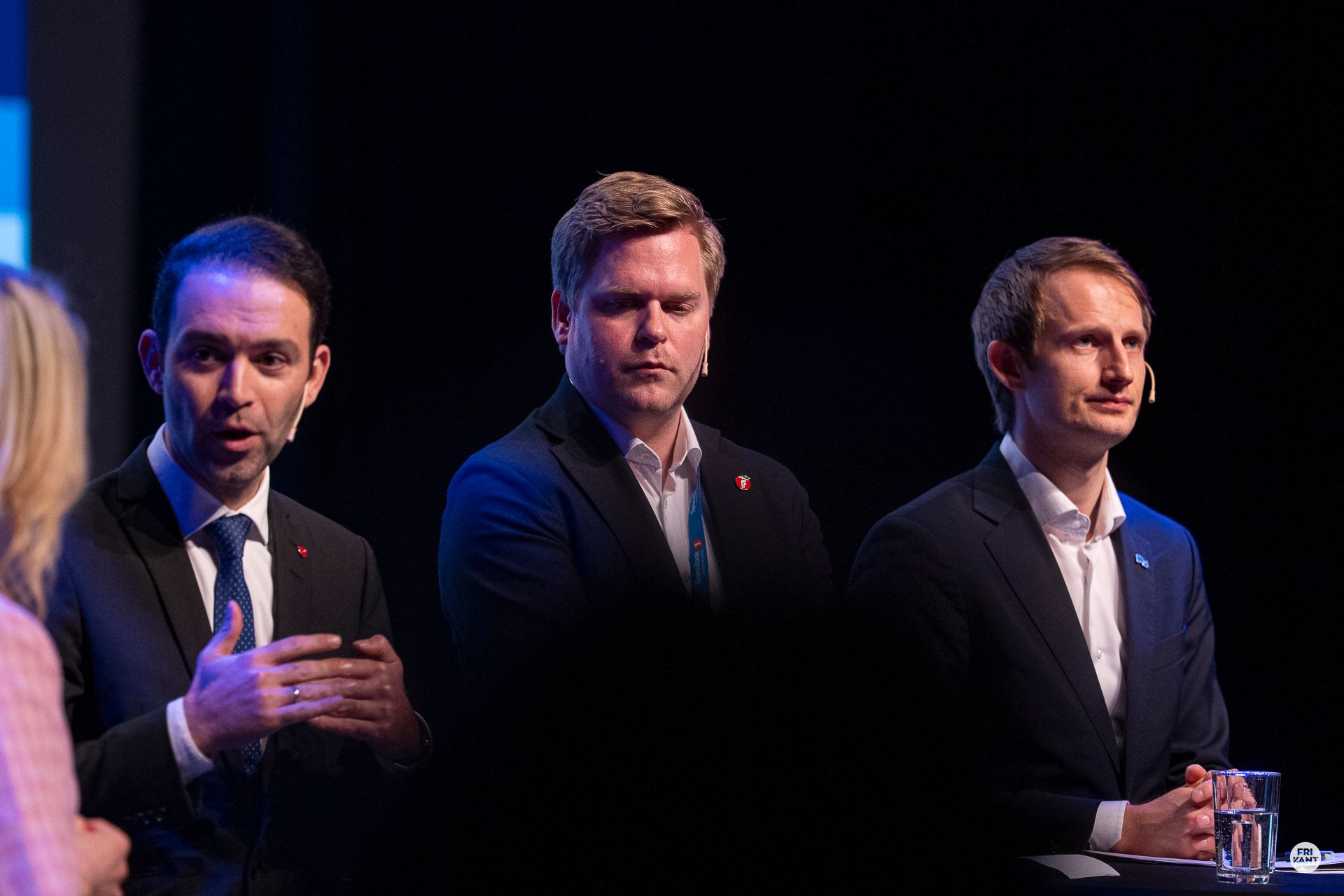 Tre menn i dress med mørk bakgrunn. Den nærmeste snakker til en lyshåret kvinnelig ordstyrer vi såvidt ser ryggen til.