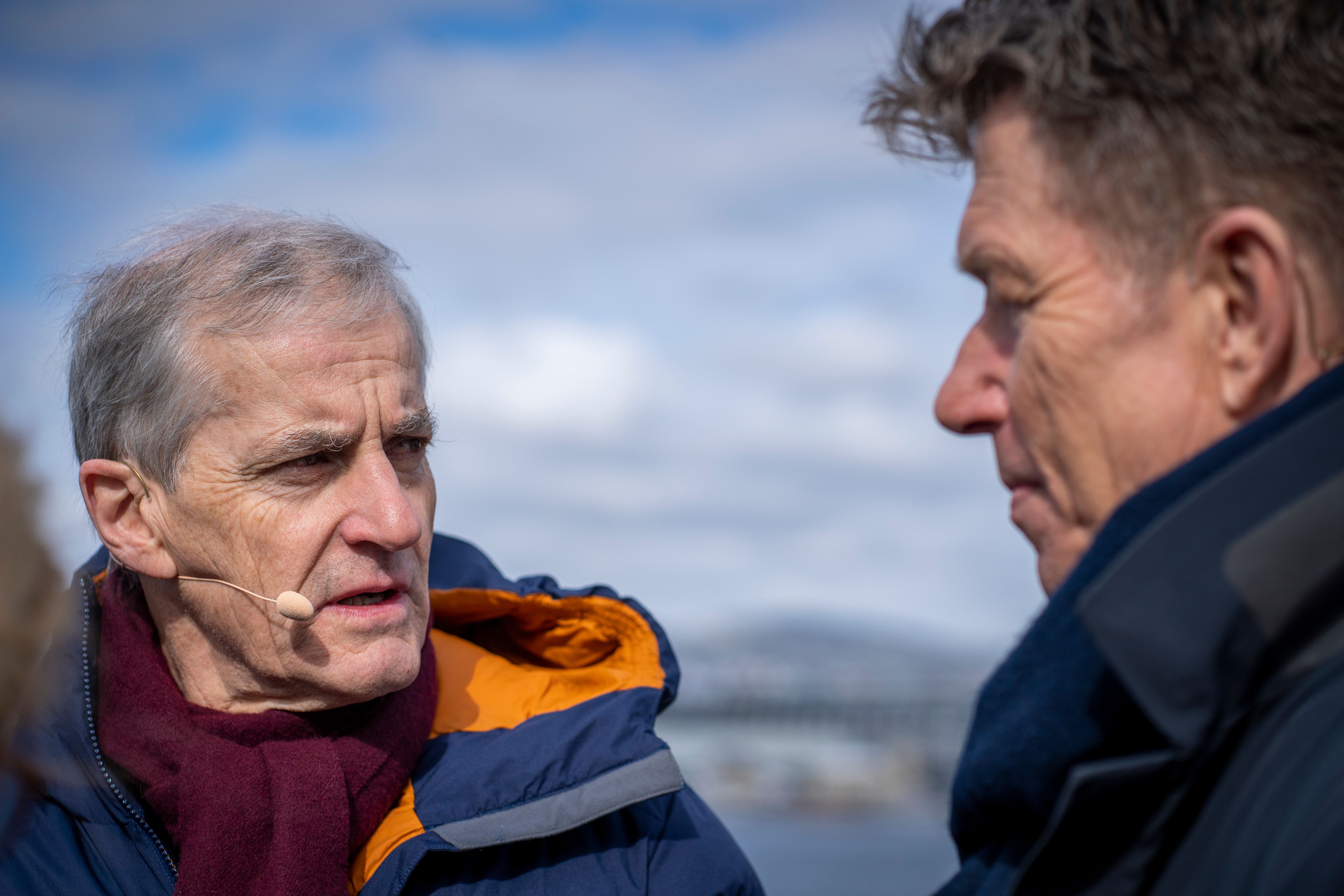 En gråhåret mann med blå jakke og vinrød skjerf snakker i en myggmikrofon til en annen mann sett i profil, begge utendørs.