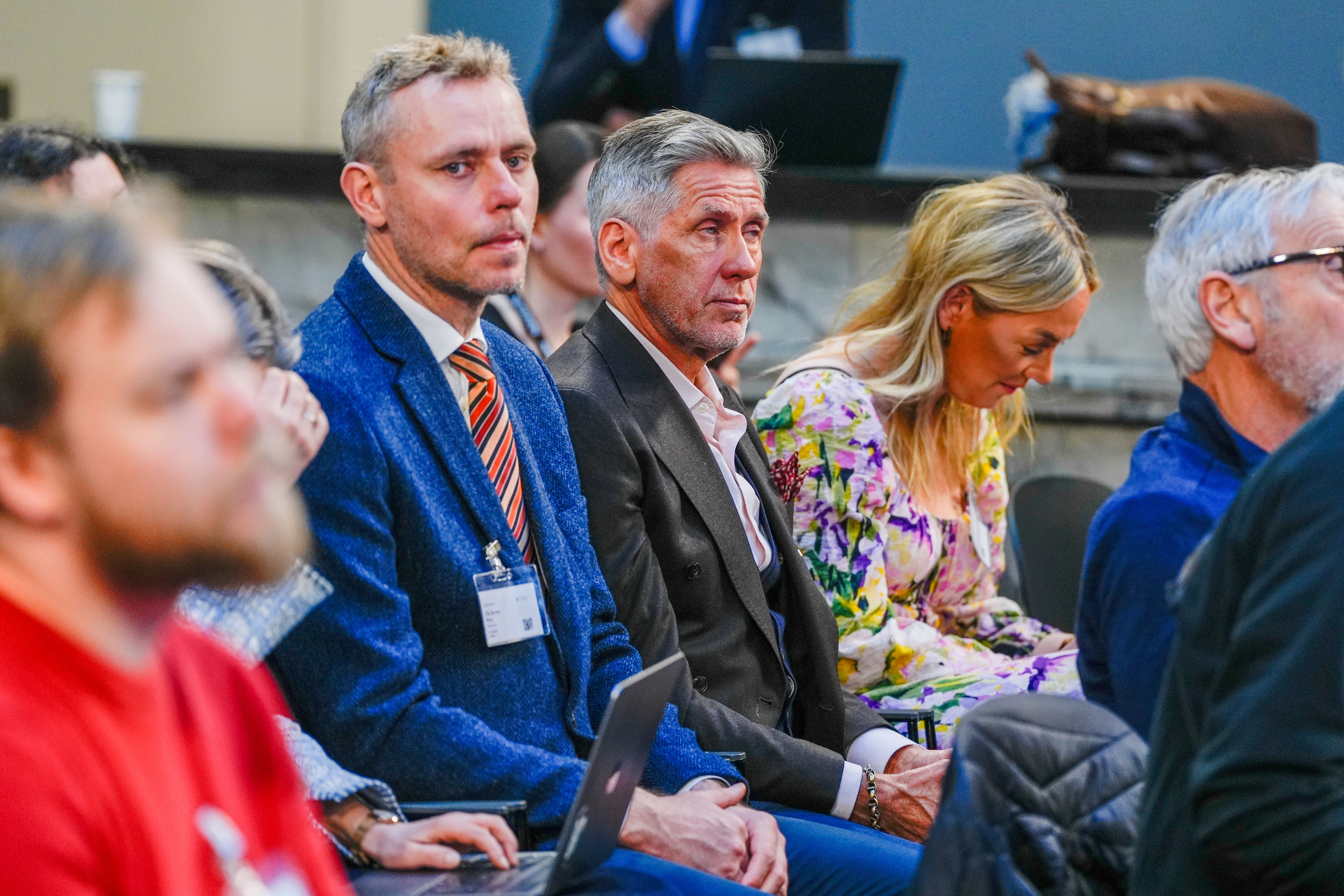 Tre deltakere på en konferanse: en mann i blå tweedjakke og slips, en eldre mann i dress og en kvinne i blomstrete kjole som ser ned.