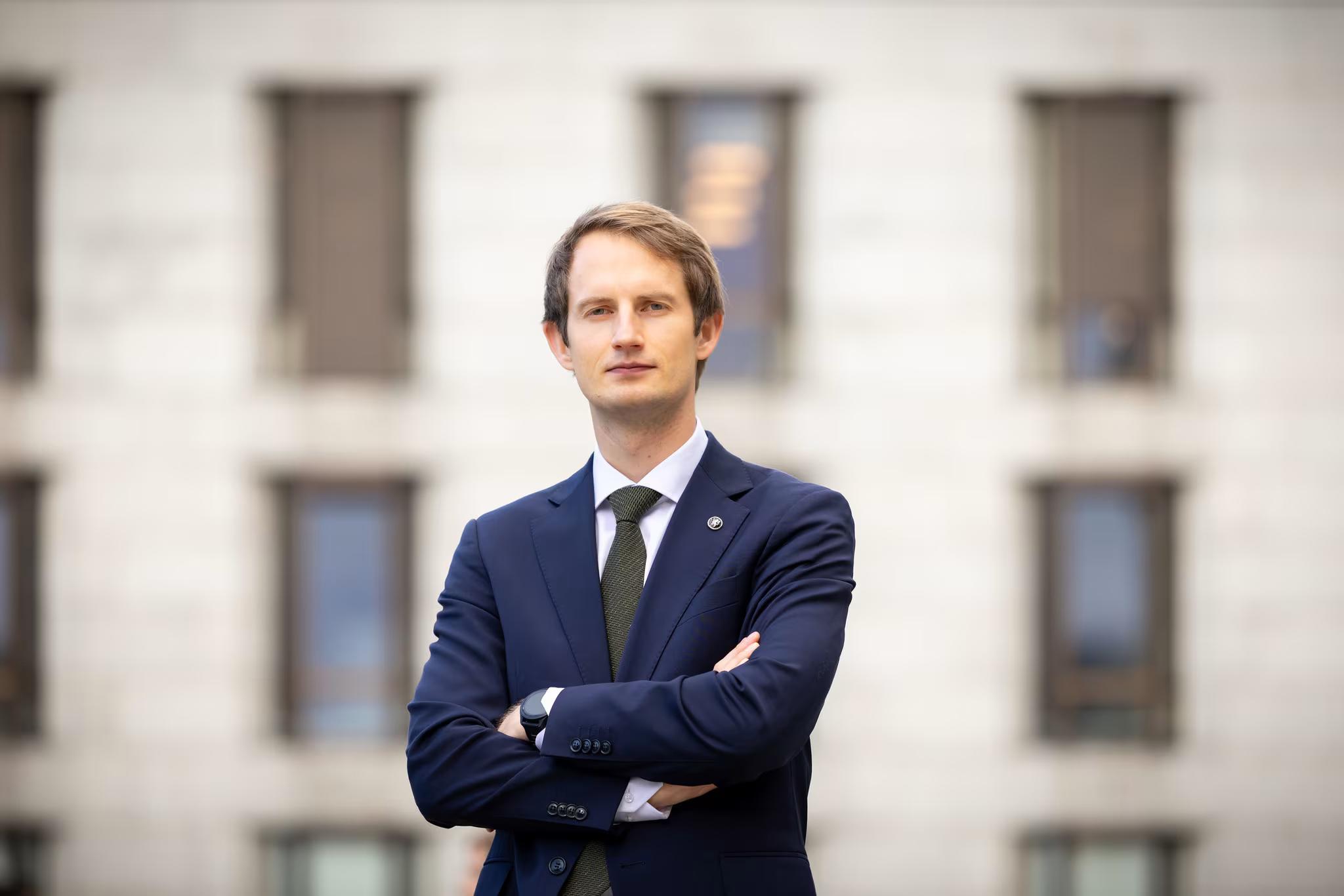 En mann i mørk dress og grønt slips med armene i kors, som ser fremover, mot en uklar bygning i bakgrunnen.