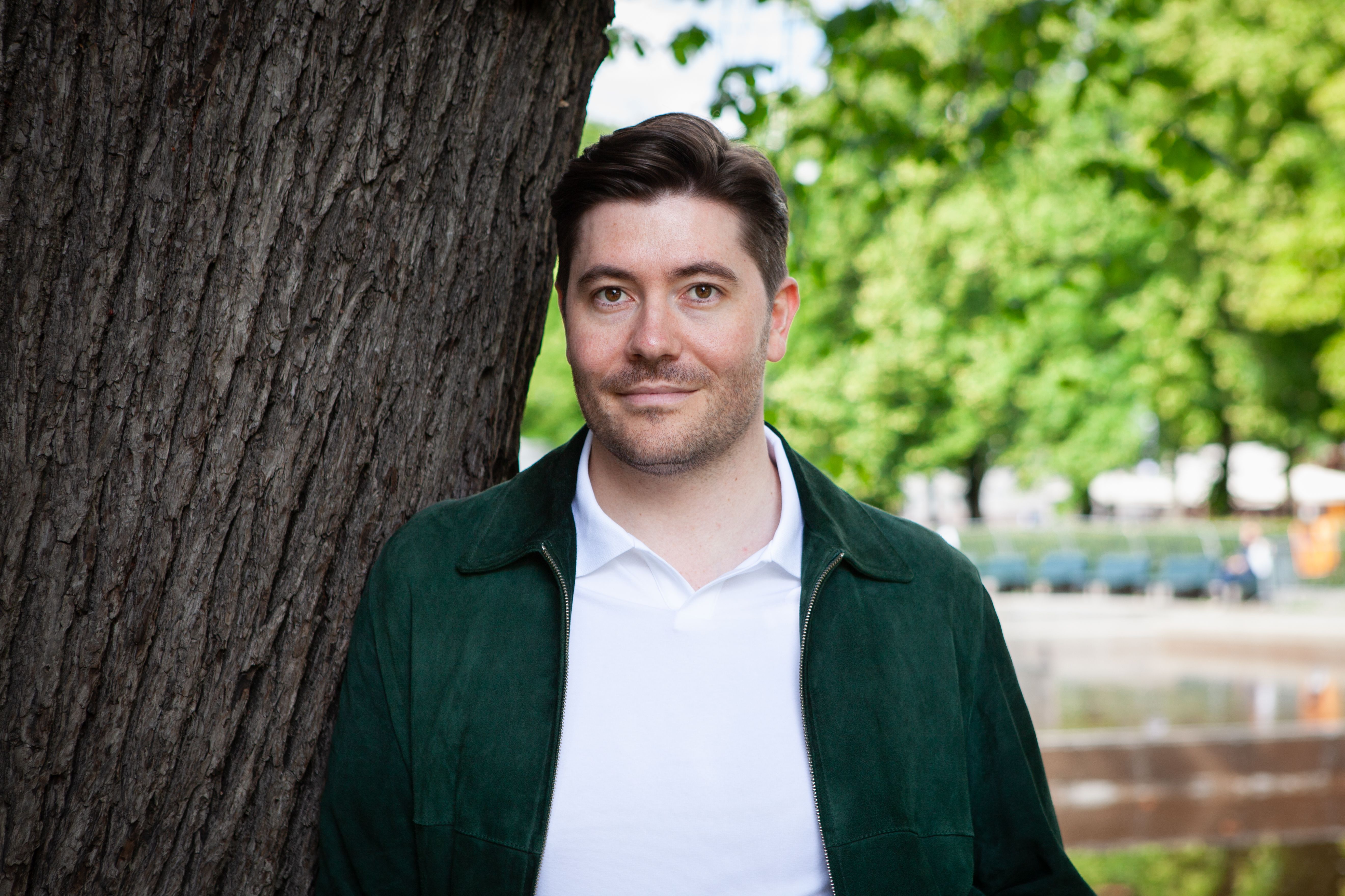 En mann i mørkegrønn jakke og hvit poloskjorte smiler mens han står ved siden av et tre, med en uklar park i bakgrunnen.