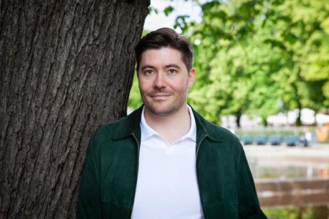 En mann i mørkegrønn jakke og hvit poloskjorte smiler mens han står ved siden av et tre, med en uklar park i bakgrunnen.