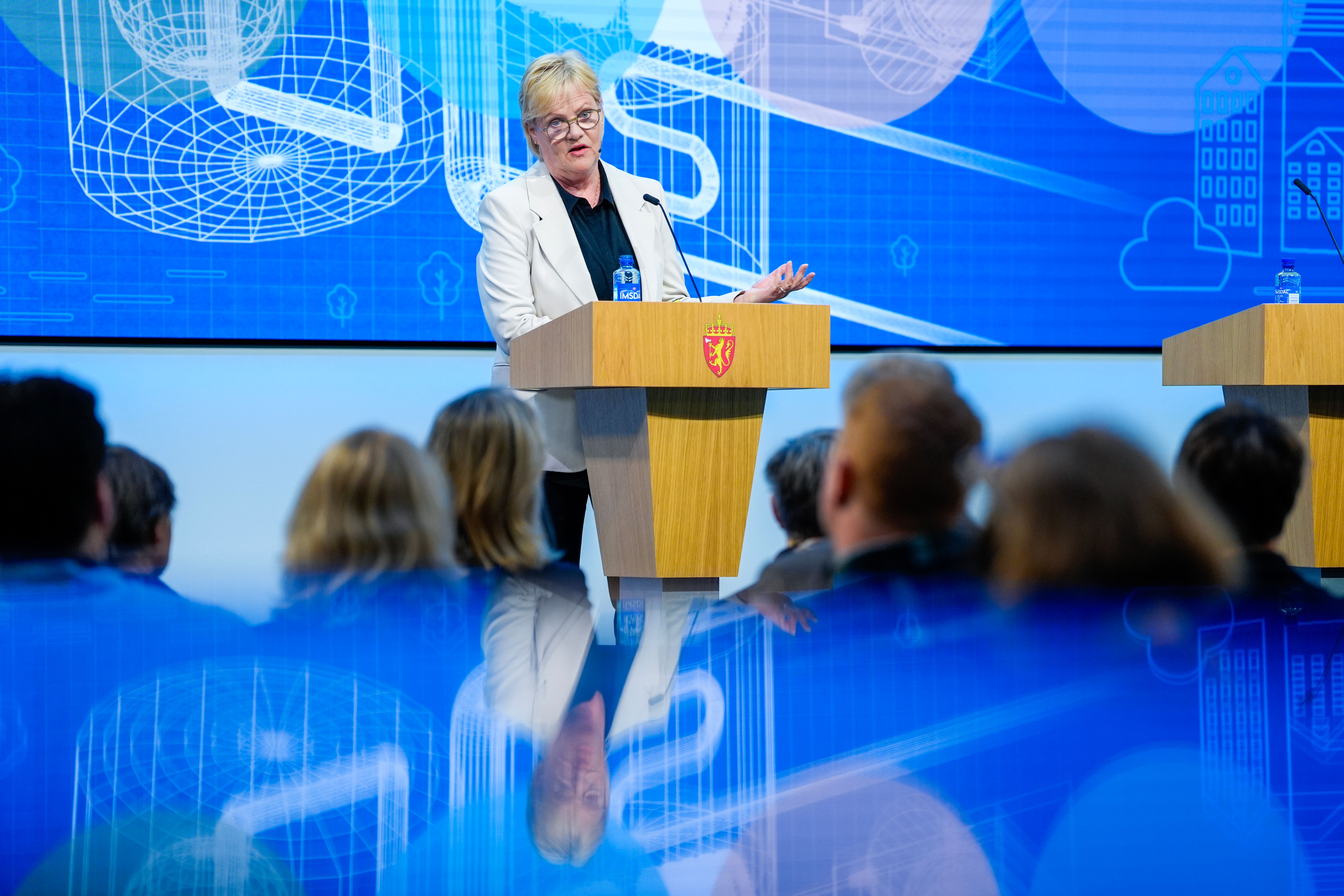 En kvinne i hvit blazer taler fra en talerstol med et statlig våpenskjold, mot en blå digital bakgrunn, med et publikum uskarpt i forgrunnen.