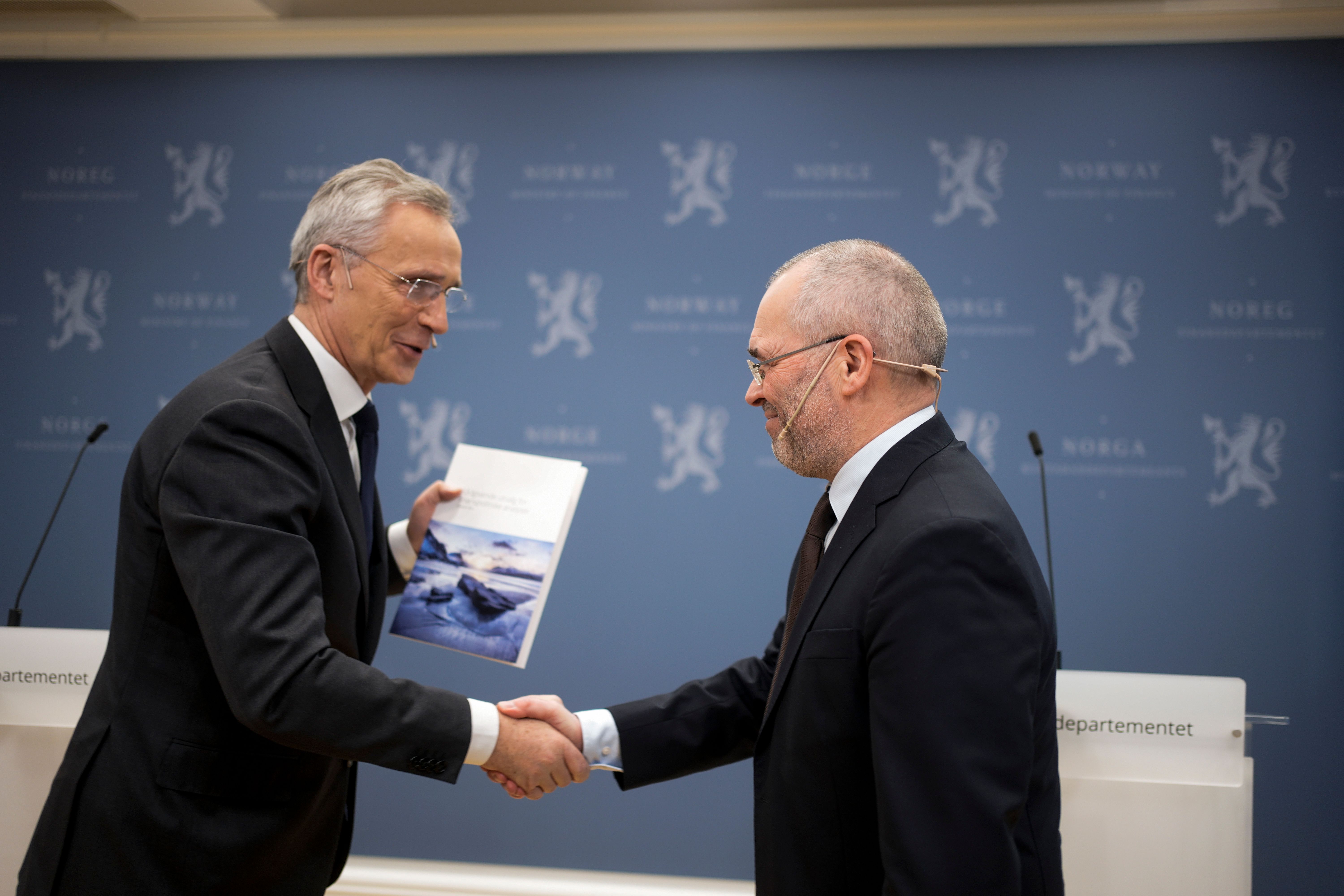 Finansminister Jens Stoltenberg og utvalgsleder Ragnar Torvik, begge i dress, tar hverandre i hånden, Stoltenberg holder et dokument, foran en blå vegg med Norges riksvåpen.