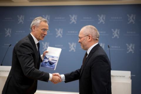 Finansminister Jens Stoltenberg og utvalgsleder Ragnar Torvik, begge i dress, tar hverandre i hånden, Stoltenberg holder et dokument, foran en blå vegg med Norges riksvåpen.