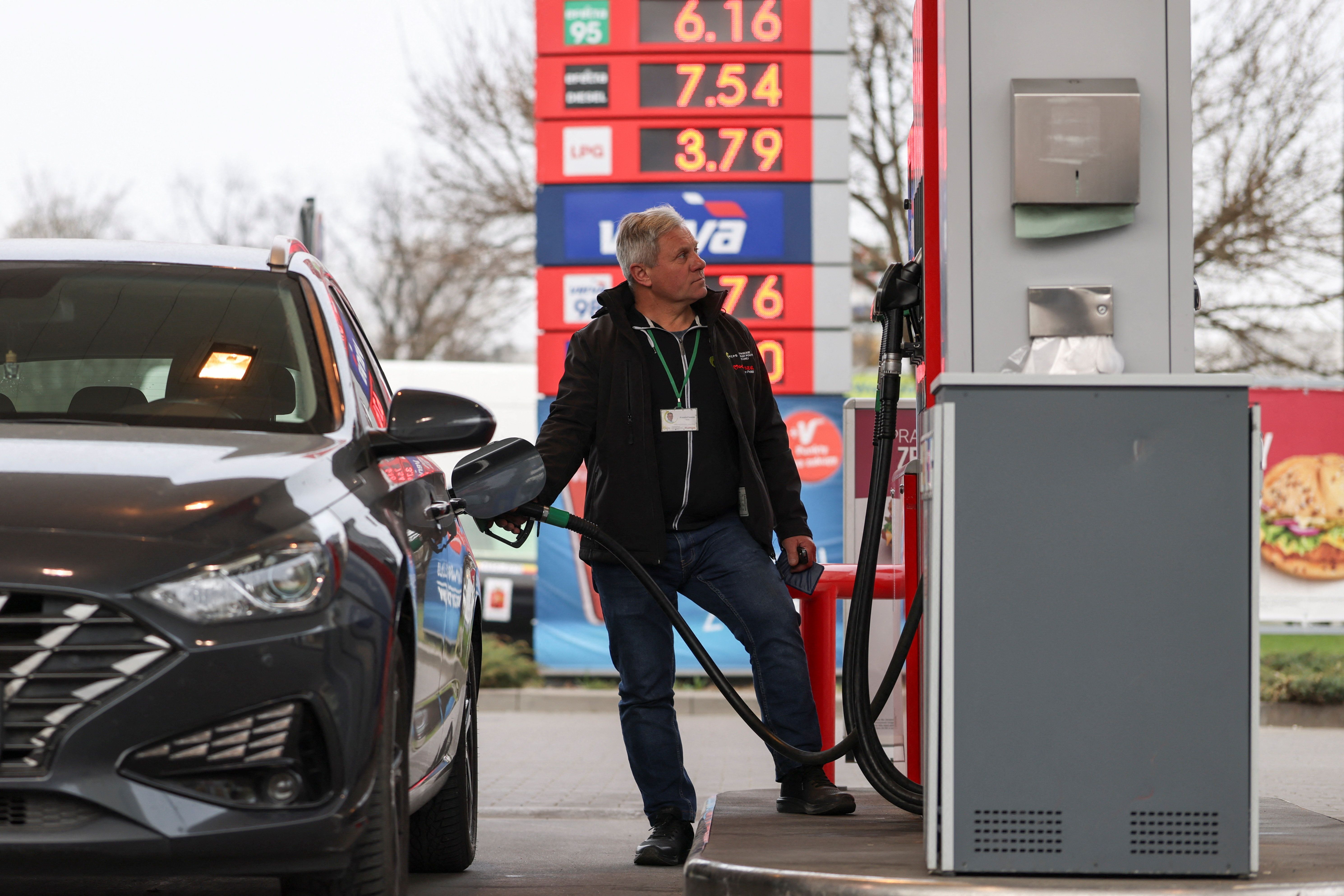 A man fuels a gray car at a gas station, looking up at the displayed fuel prices.