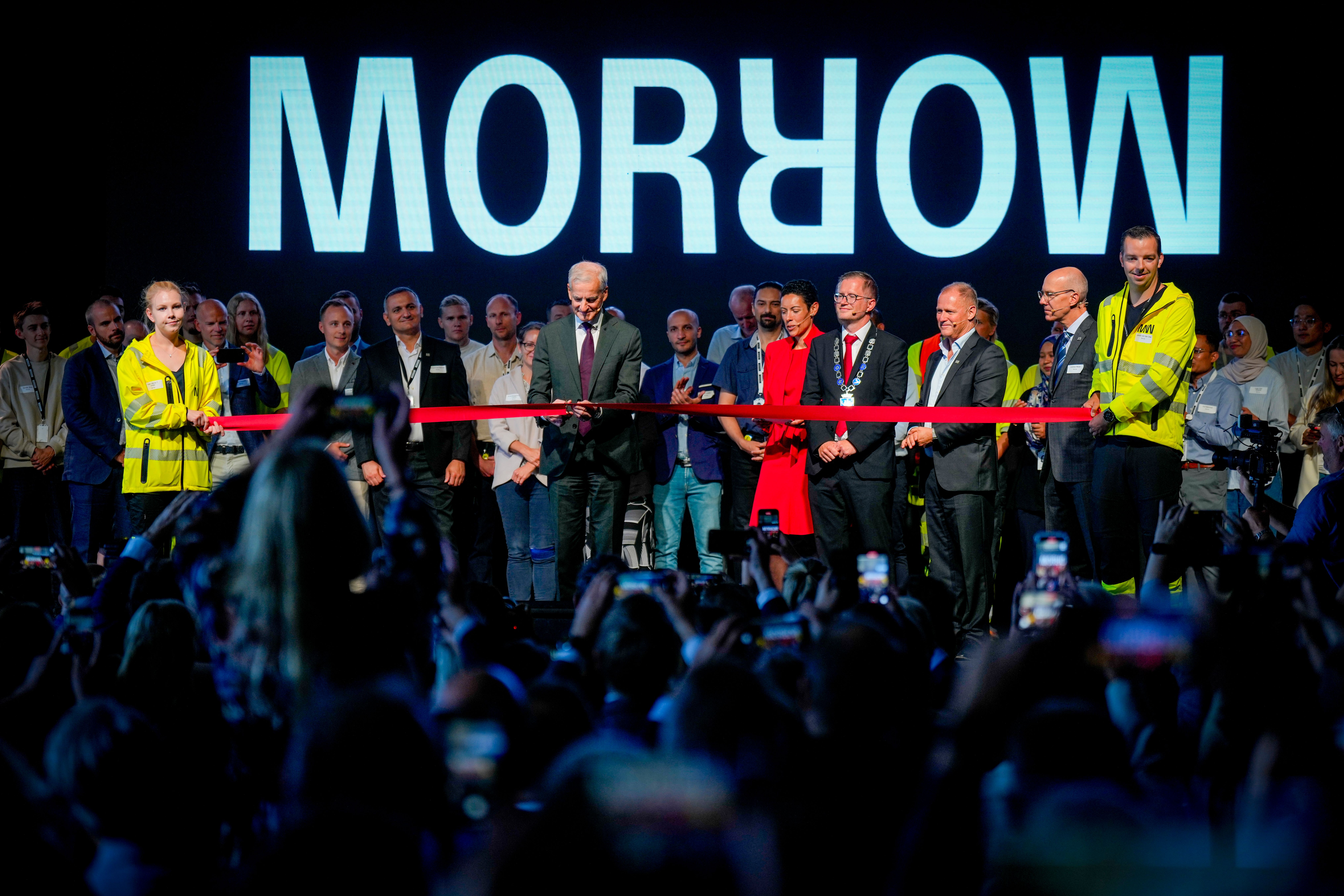 A man in a suit cuts a red ribbon at a Morrow ceremony, with officials and a crowd watching.