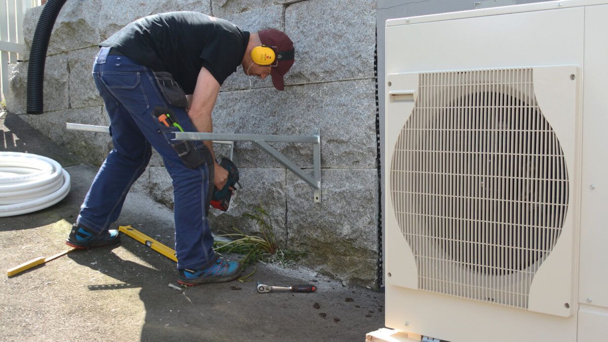 En tekniker med hørselvern borer i en steinvegg for å montere en utendørs varmepumpe-enhet.