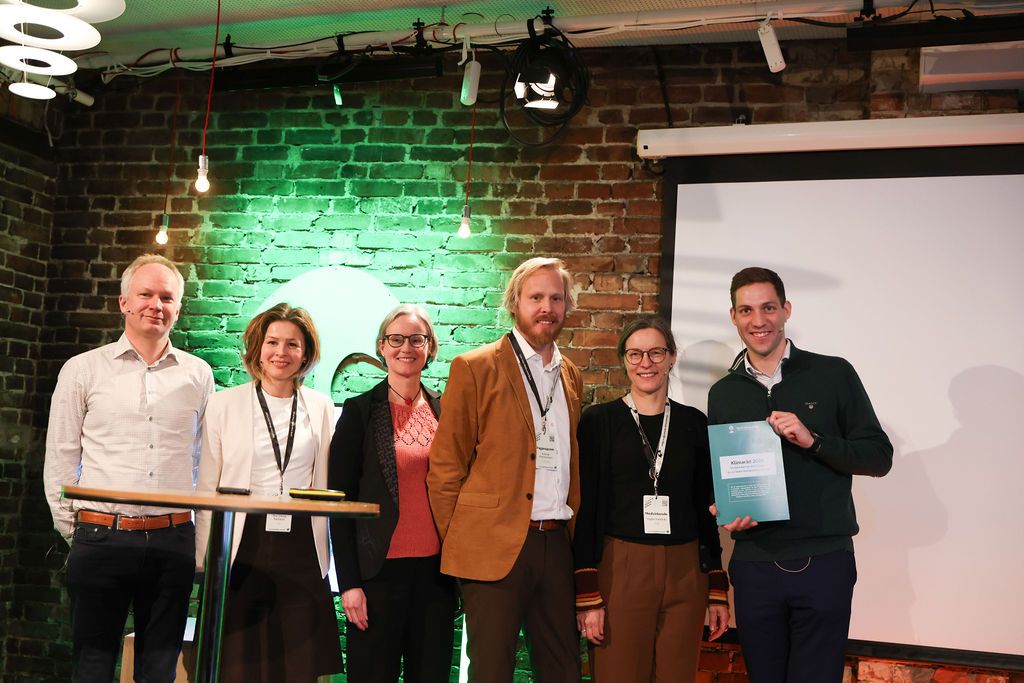 Seks personer står og smiler, med en mann til høyre som holder et dokument.