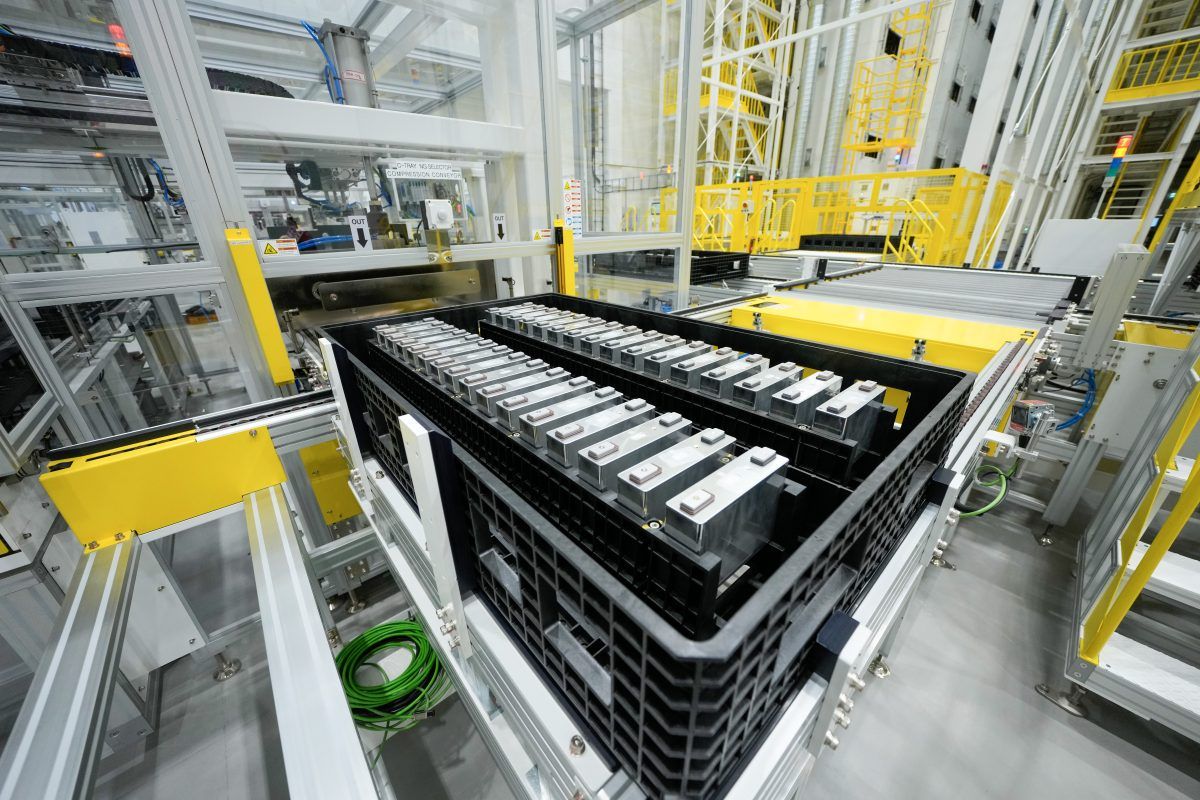 En svart plastbeholder rommer flere rektangulære battericeller i et industrianlegg med metallkonstruksjoner og gule sikkerhetsskinner.