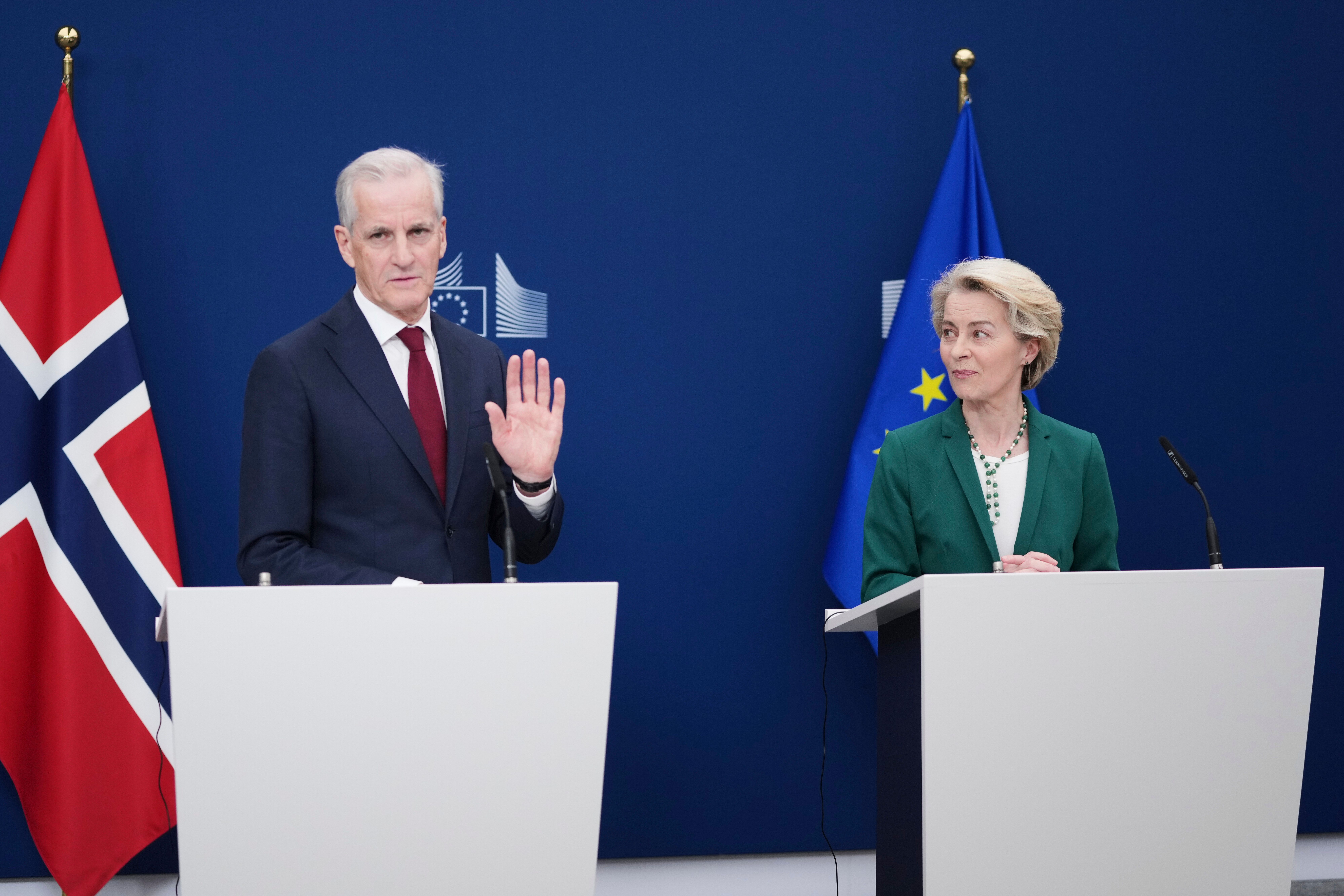 En mann i mørk dress med hånden hevet og en kvinne i grønn blazer står ved hvert sitt podium, flankert av norske og EU-flagg.