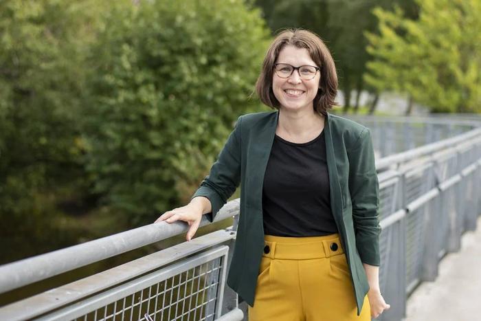 En kvinne med briller, mørkegrønn blazer og gule bukser står smilende på en bro med grønne planter i bakgrunnen.