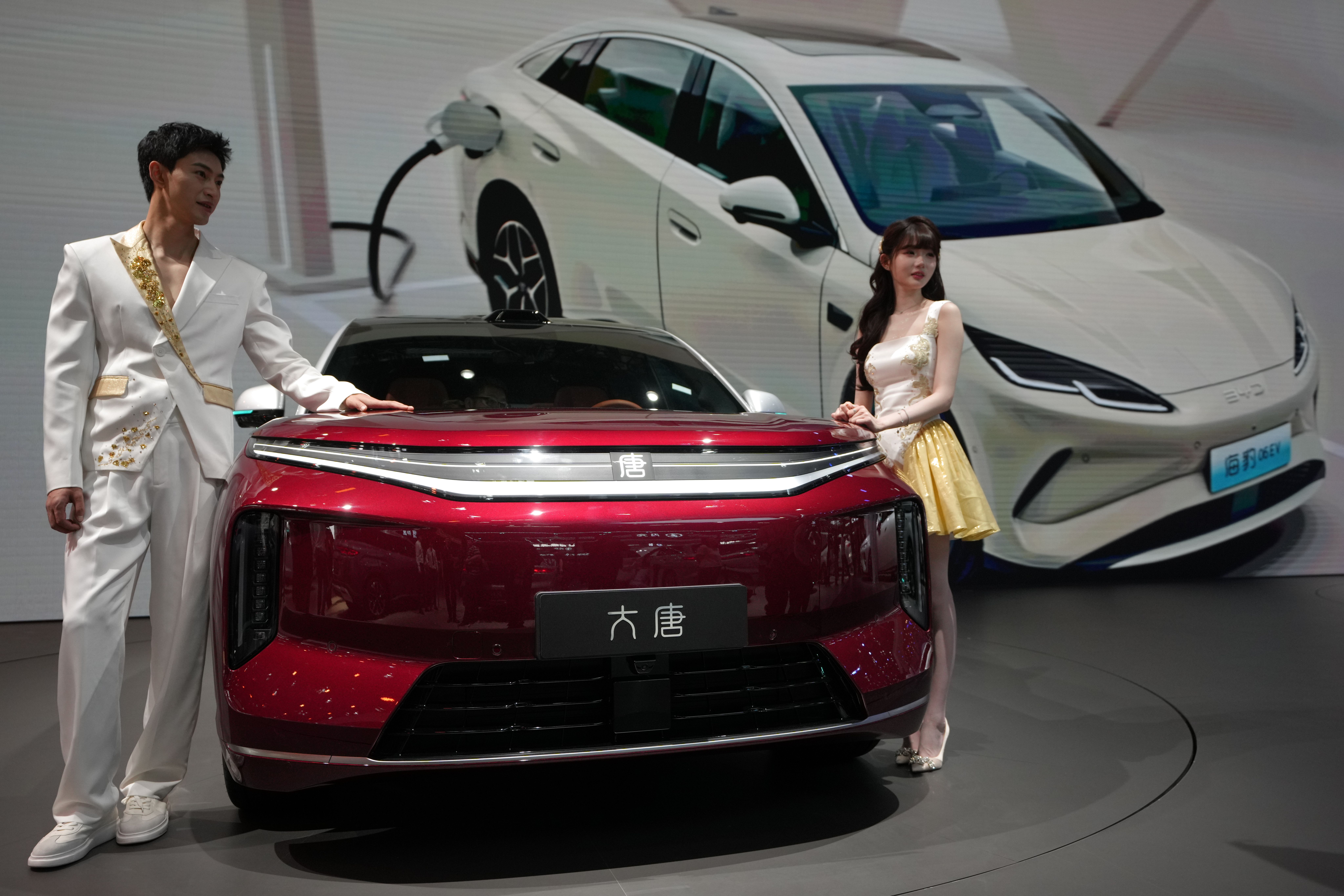 To modeller poserer ved en rød bil på en bilutstilling, med en hvit elbil som lader i bakgrunnen.