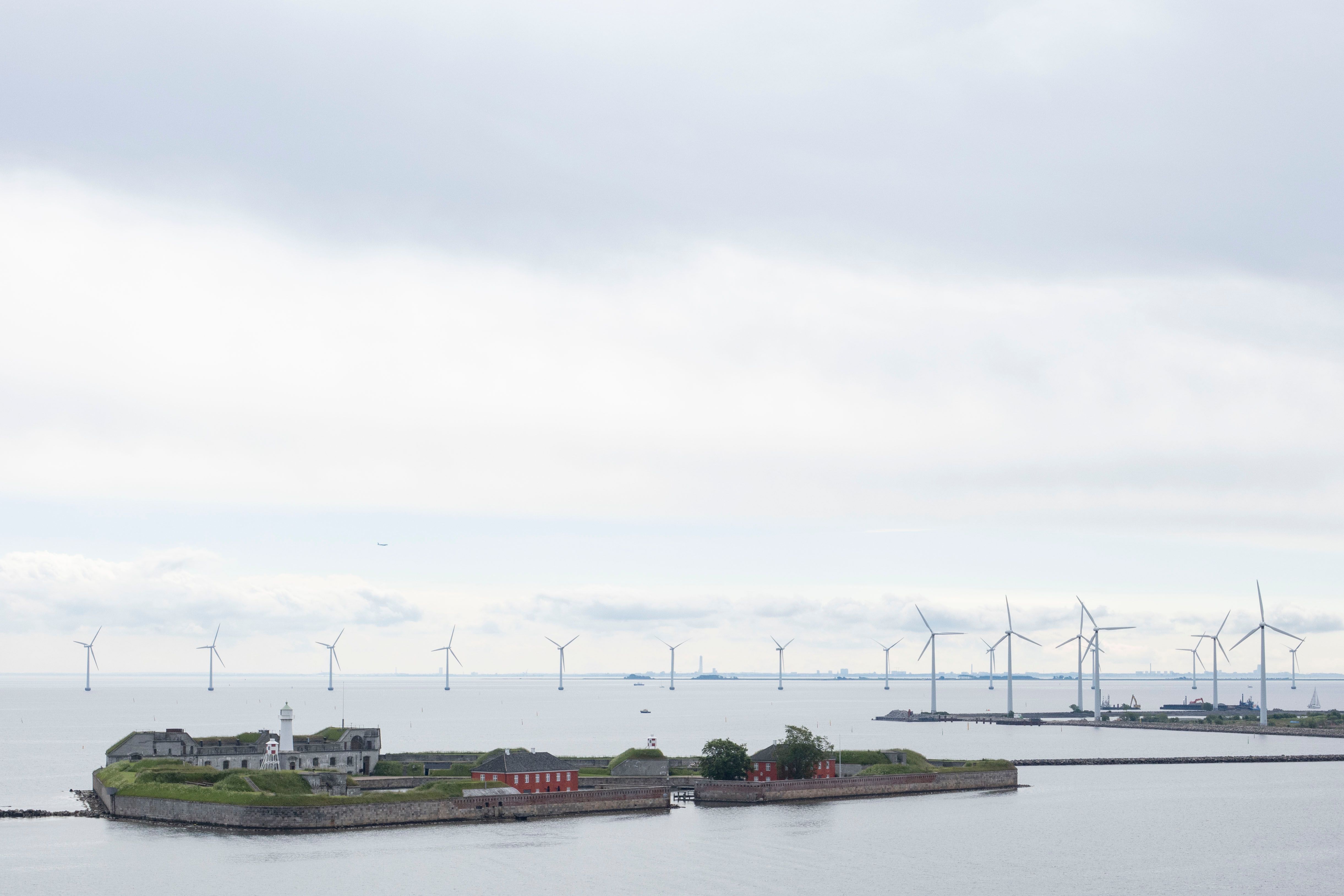 En befestet øy med et fyrtårn i forgrunnen, og en havvindpark i bakgrunnen under en skyet himmel.