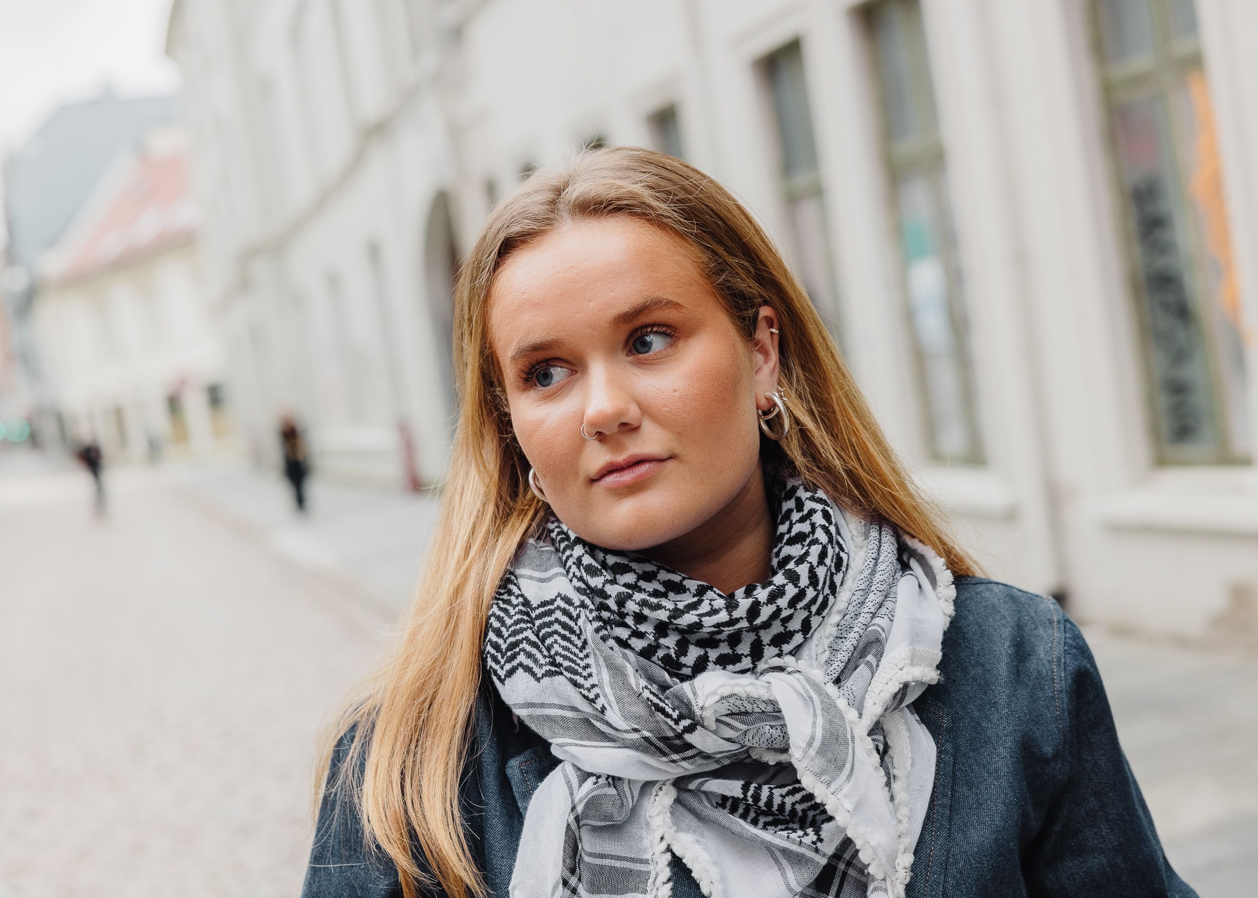 En ung blond kvinne med et svart‑hvitt mønstret skjerf ser til siden i en bygate.