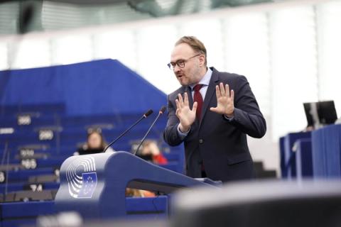 En mann i dress og slips taler fra et podium med mikrofoner i en stor forsamlingssal, mens han gestikulerer med begge hendene hevet.