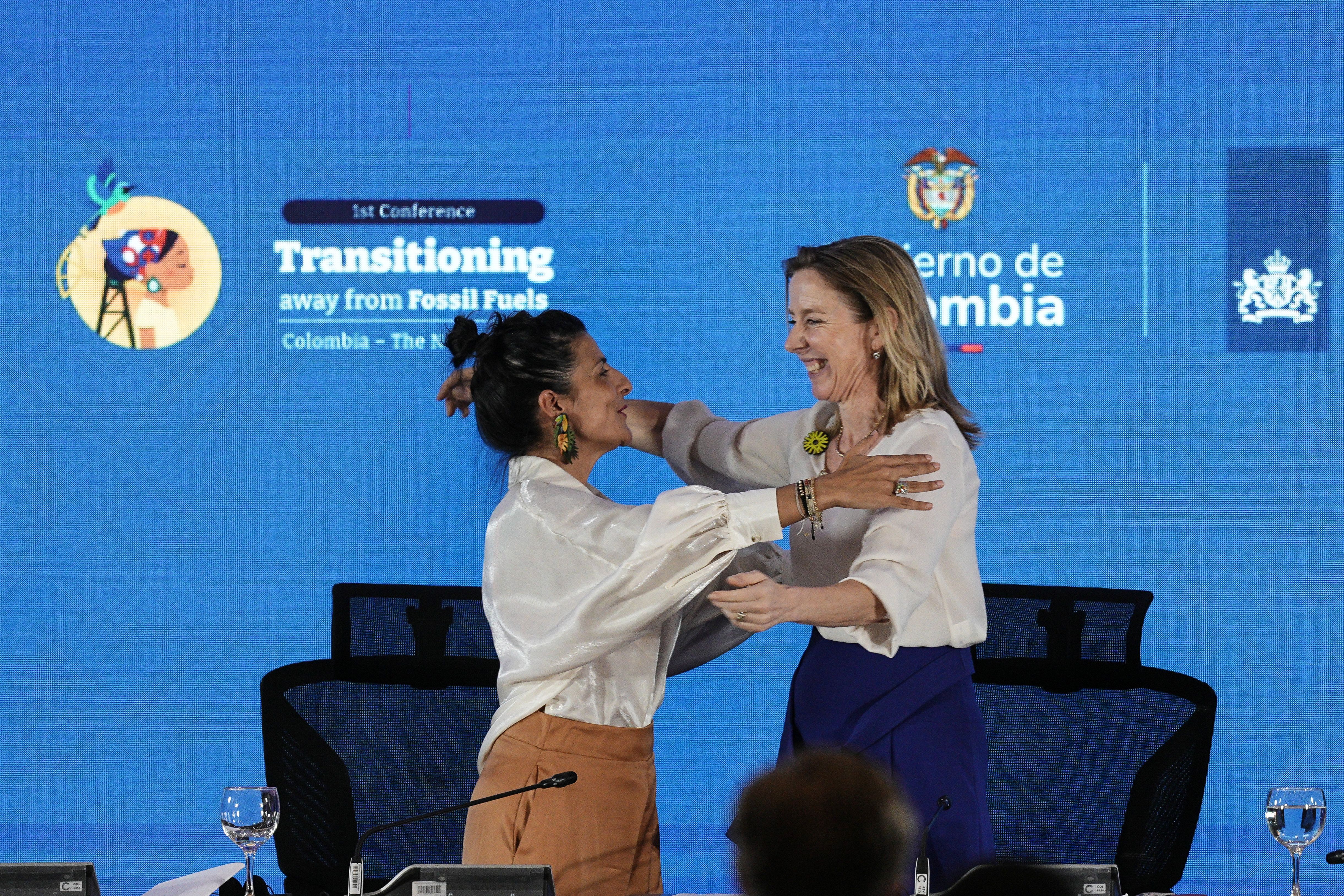 To smilende kvinner omfavner hverandre på en scene foran en blå skjerm med teksten «Transitioning away from Fossil Fuels.»