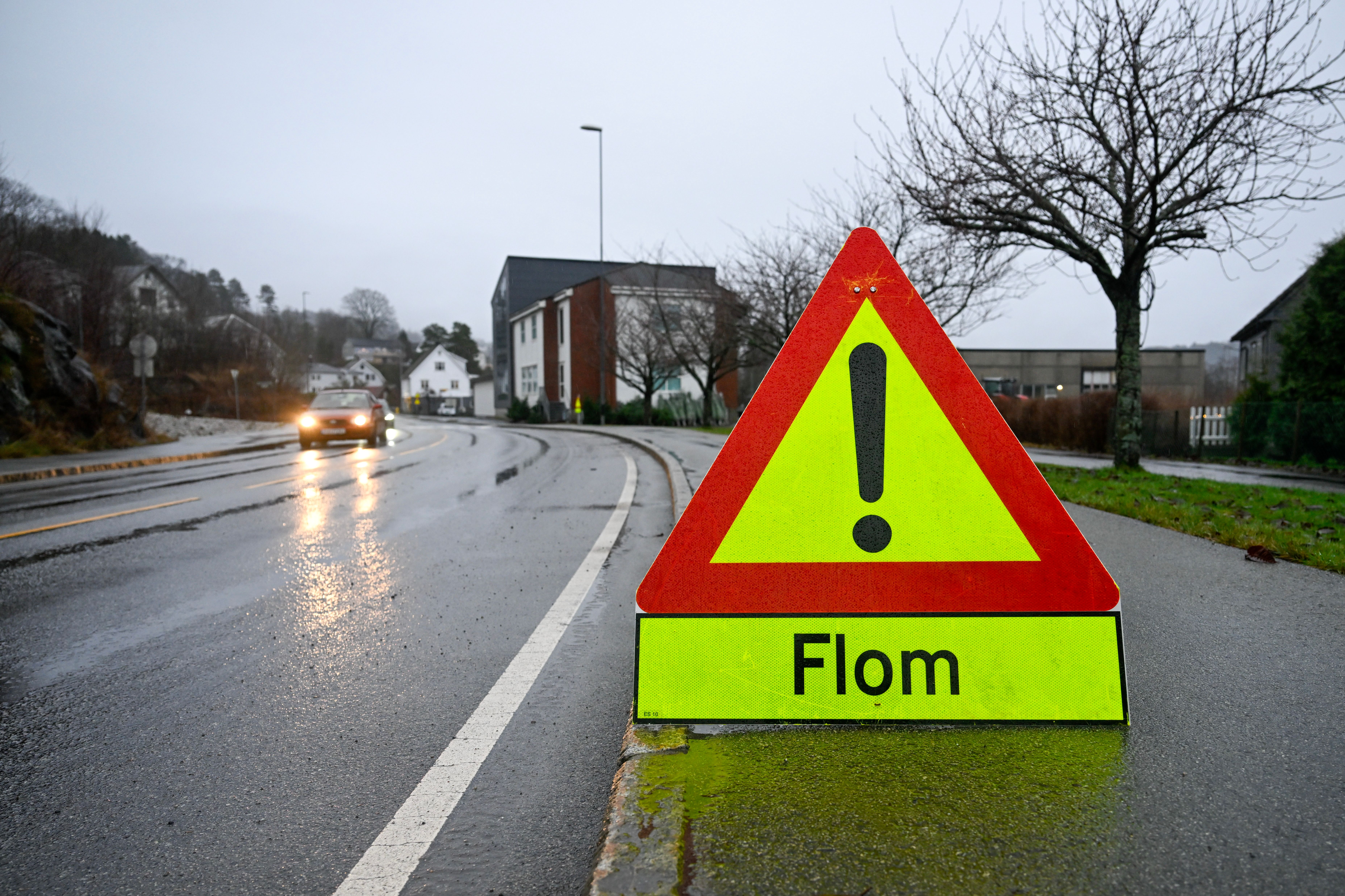 Et gult og rødt varselskilt med et utropstegn og teksten «Flom» står ved siden av en våt vei, mens en bil nærmer seg i det fjerne.