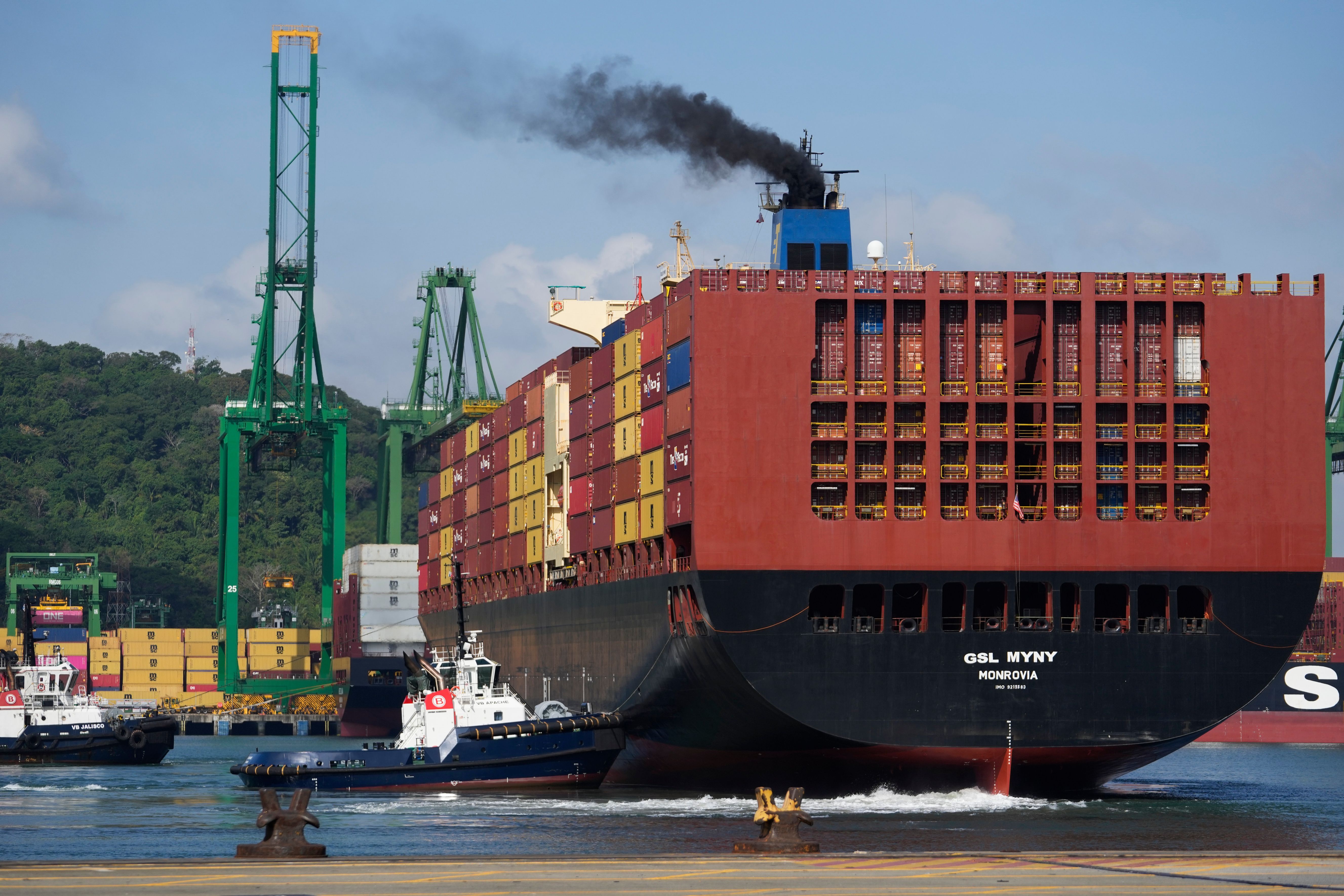 Et stort containerskip, GSL MYNY MONROVIA, som slipper ut mørk røyk, blir assistert av to taubåter i en havn med kraner.