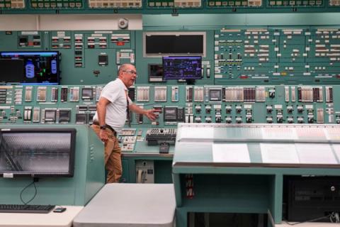 En mann gestikulerer mot et komplekst kontrollpanel i et industrielt kontrollrom.