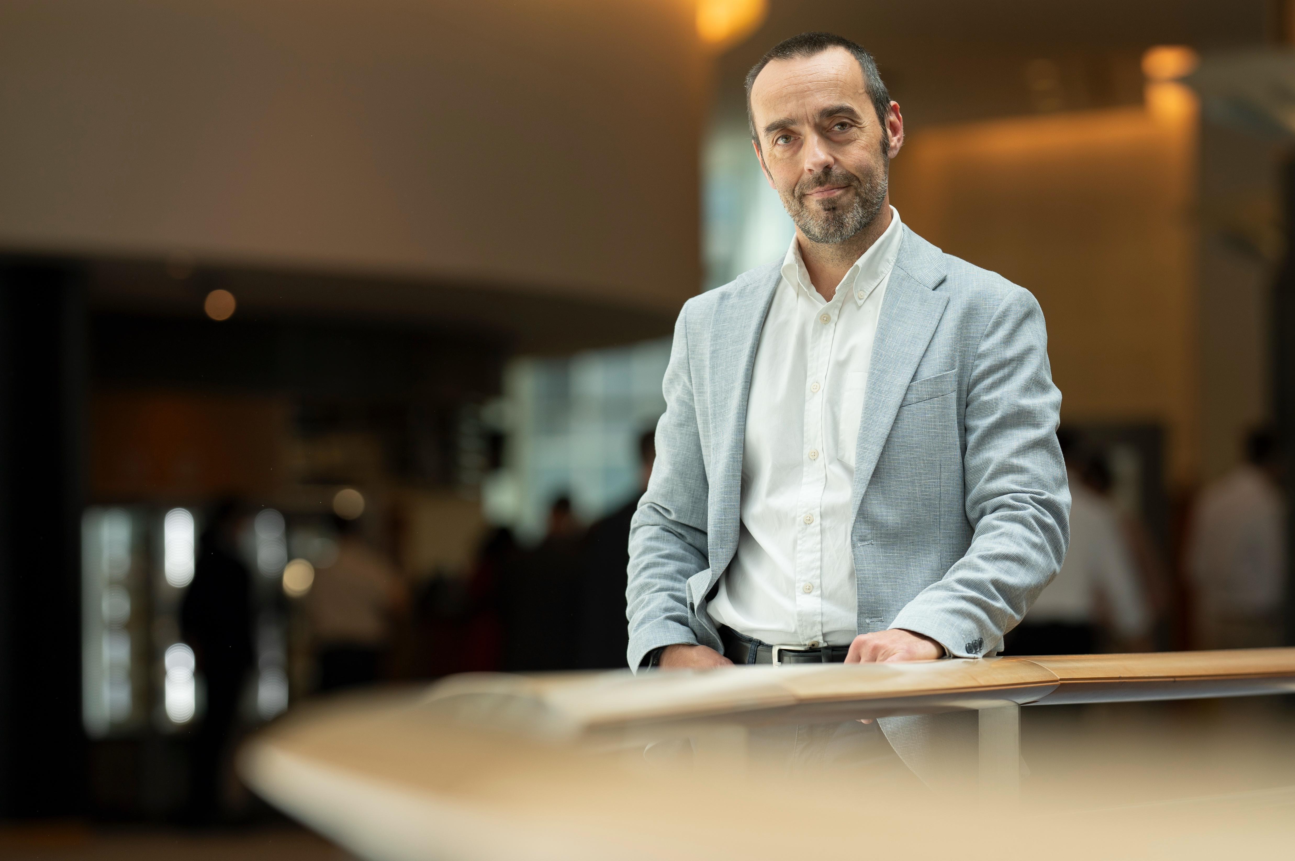 A man in a light blue blazer and white shirt leans on a wooden railing, smiling slightly at the camera in an indoor setting.