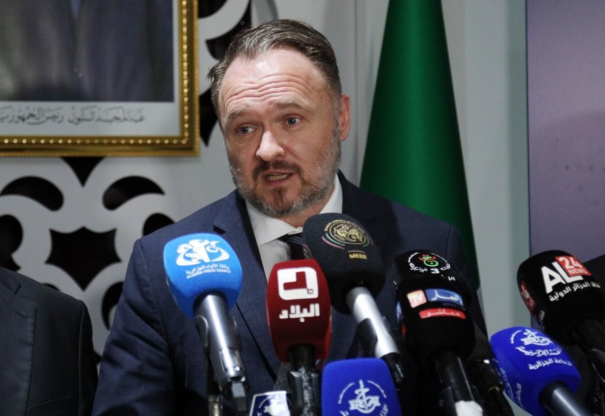 En mann i dress snakker i en rekke mikrofoner på en pressekonferanse, flankert av to andre tjenestemenn, med EU- og algeriske flagg i bakgrunnen.