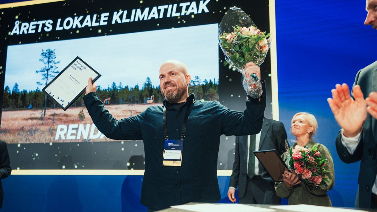 En mann med diplom og bukett står på scenen foran en storskjerm med teksten "ÅRETS LOKALE KLIMATILTAK", mens folk rundt ham applauderer.