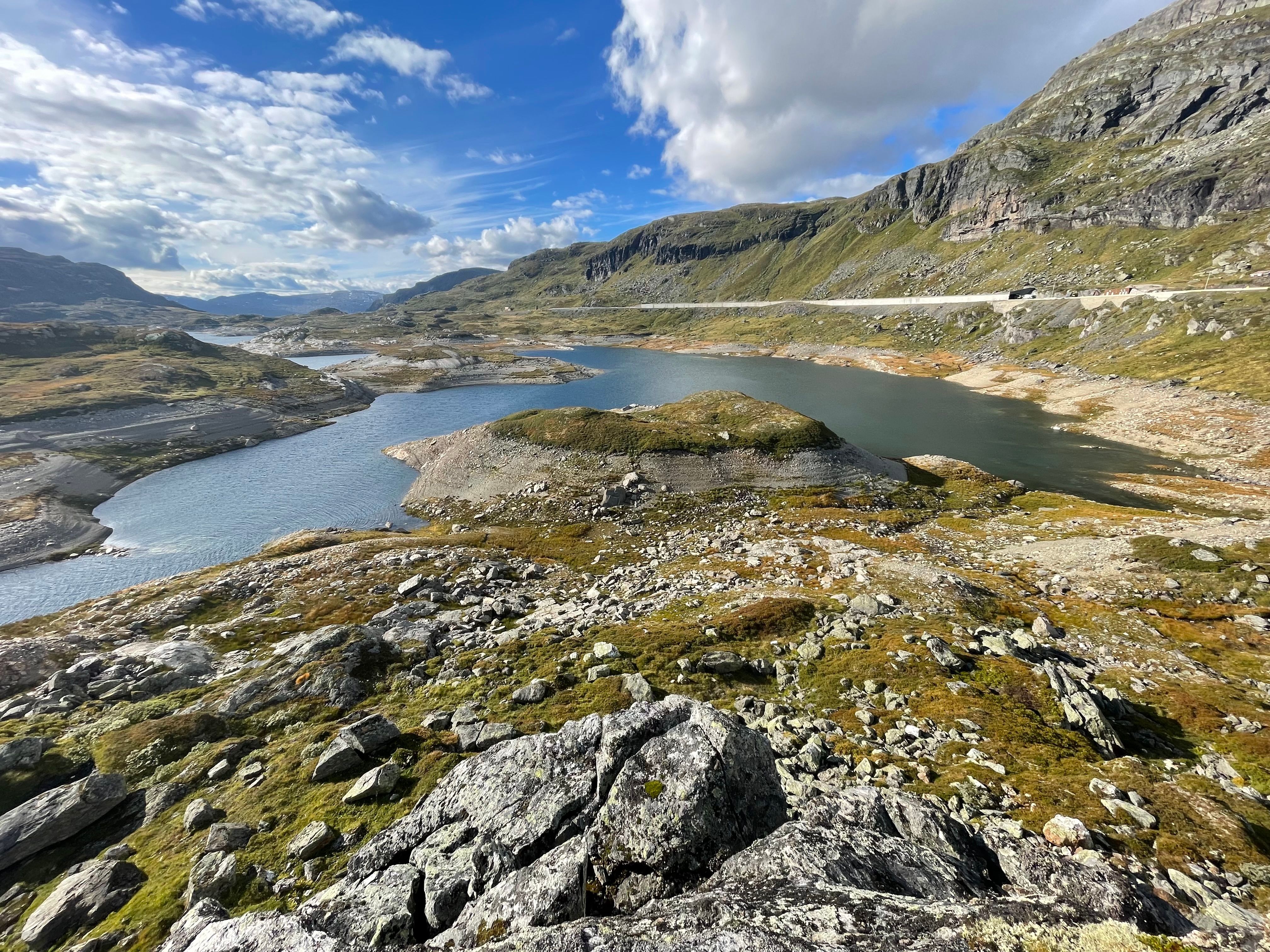 Fjellandskap med innsjøer, en svingete vei og steinete terreng under en delvis skyet himmel.
