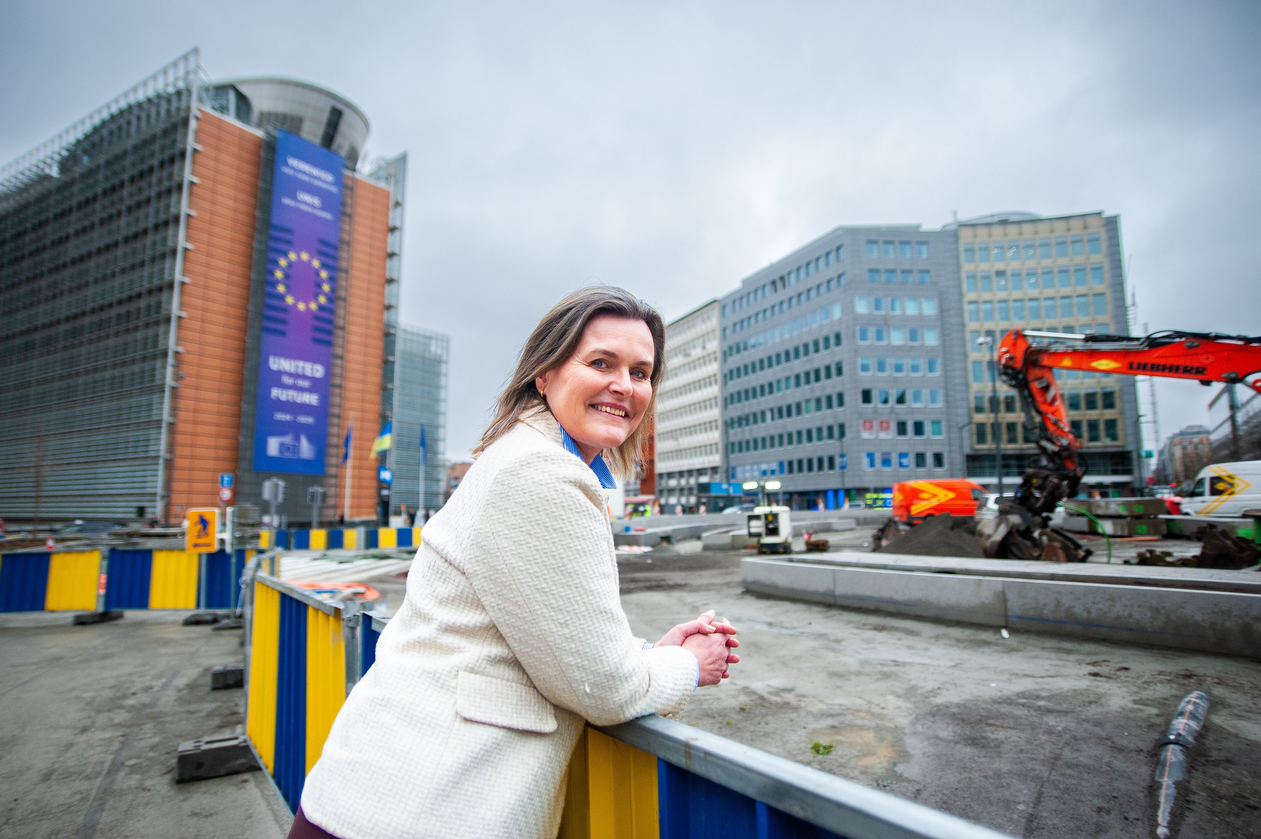 A smiling woman in a light jacket leans on a barrier, with the EU Commission building featuring a 'United for our Future' banner and a construction site behind her.