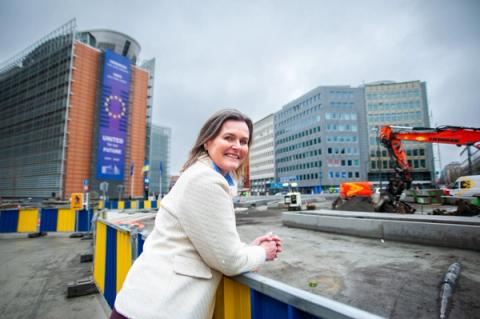 A smiling woman in a light jacket leans on a barrier, with the EU Commission building featuring a 'United for our Future' banner and a construction site behind her.