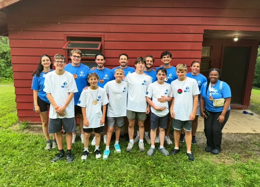 Teenagers and young adults in matching camp T-shirts pose in front of a red wooden cabin.