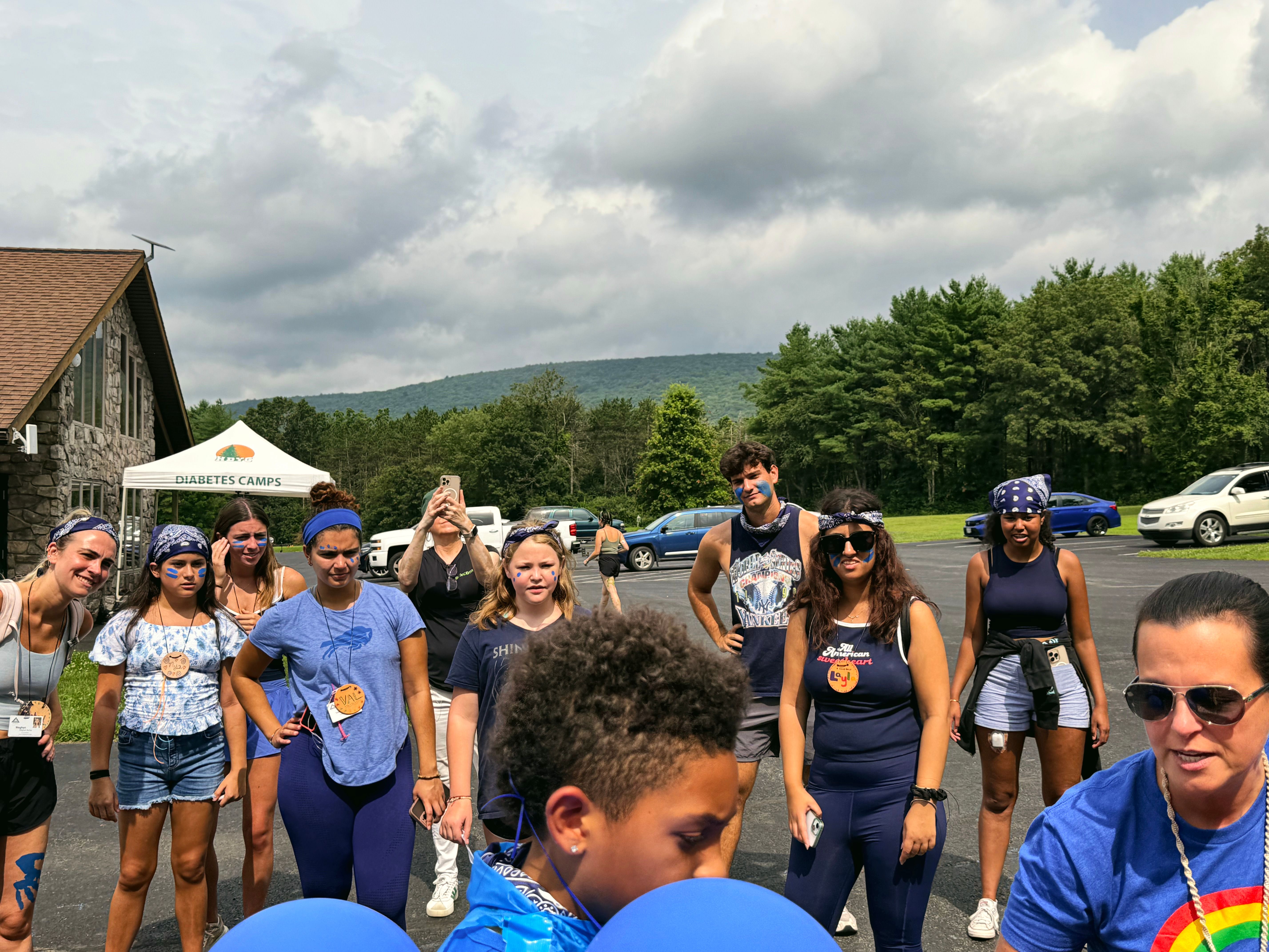 A group of young people stand outside in front of a tent that reads Diabetes Camps, with trees and mountains behind them
