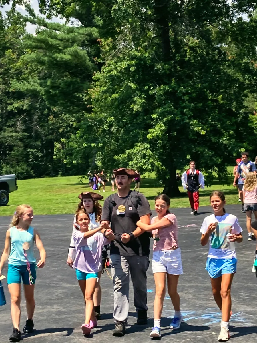 A counselor walks arm in arm with two campers on a sunny camp path