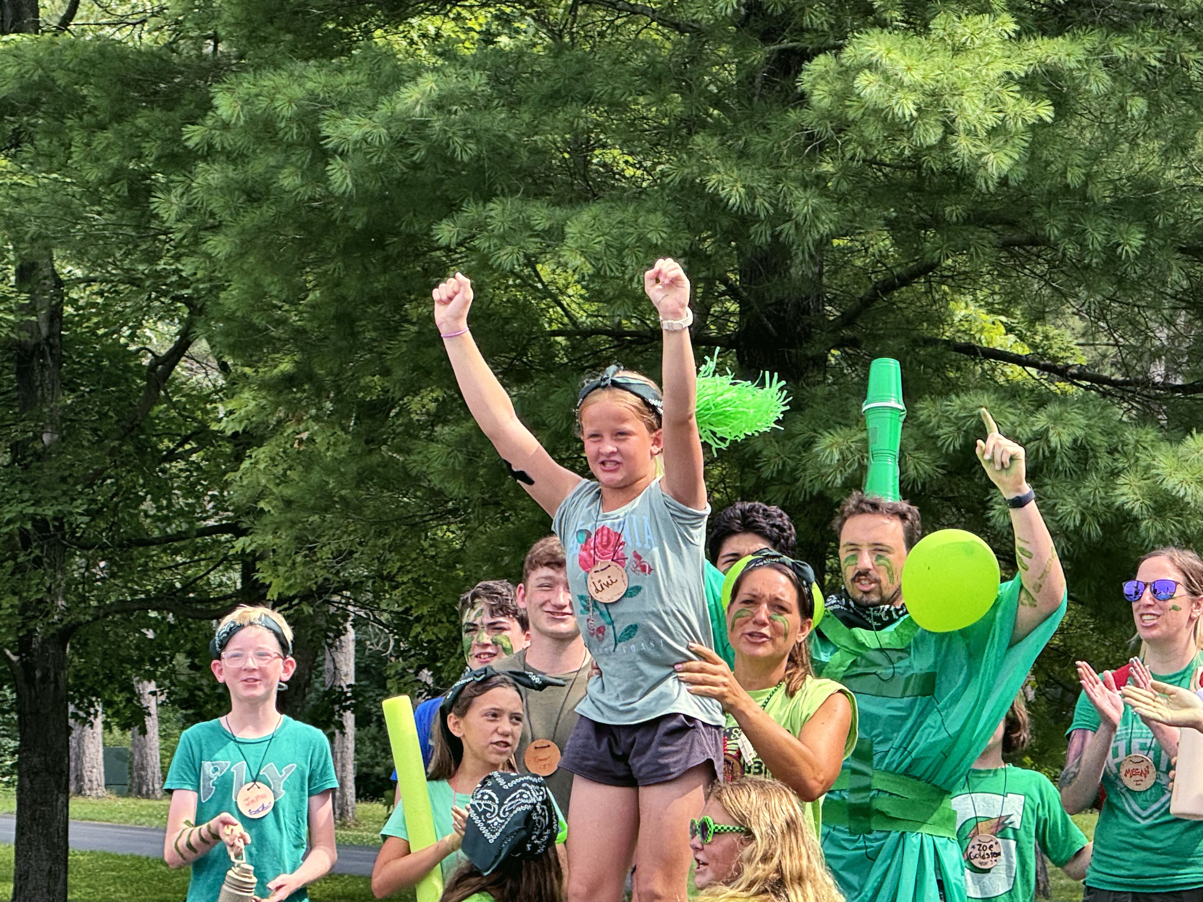 Campers and counselors in green shirts cheer with their arms in the air at camp