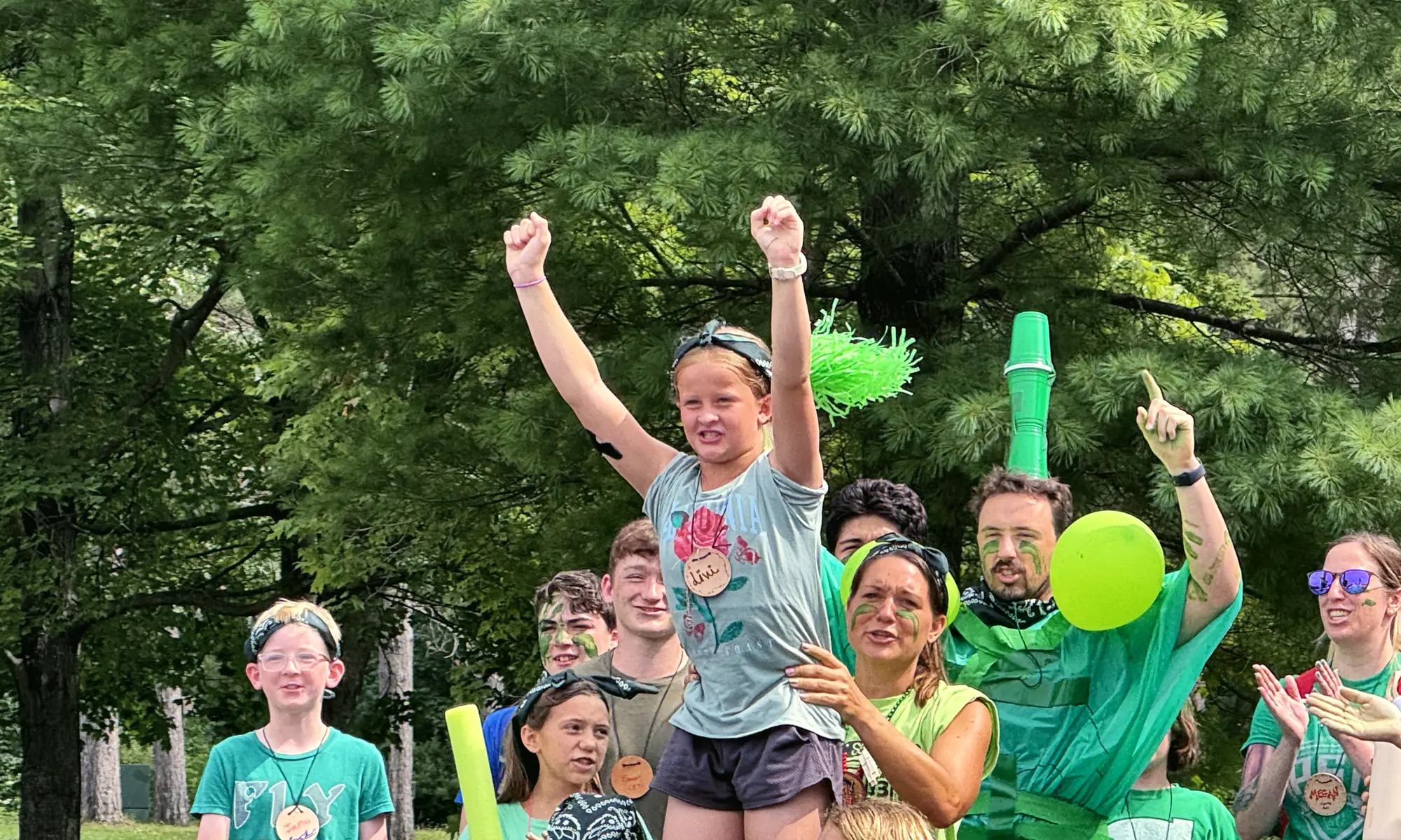 Campers and counselors in green shirts cheer with their arms in the air at camp