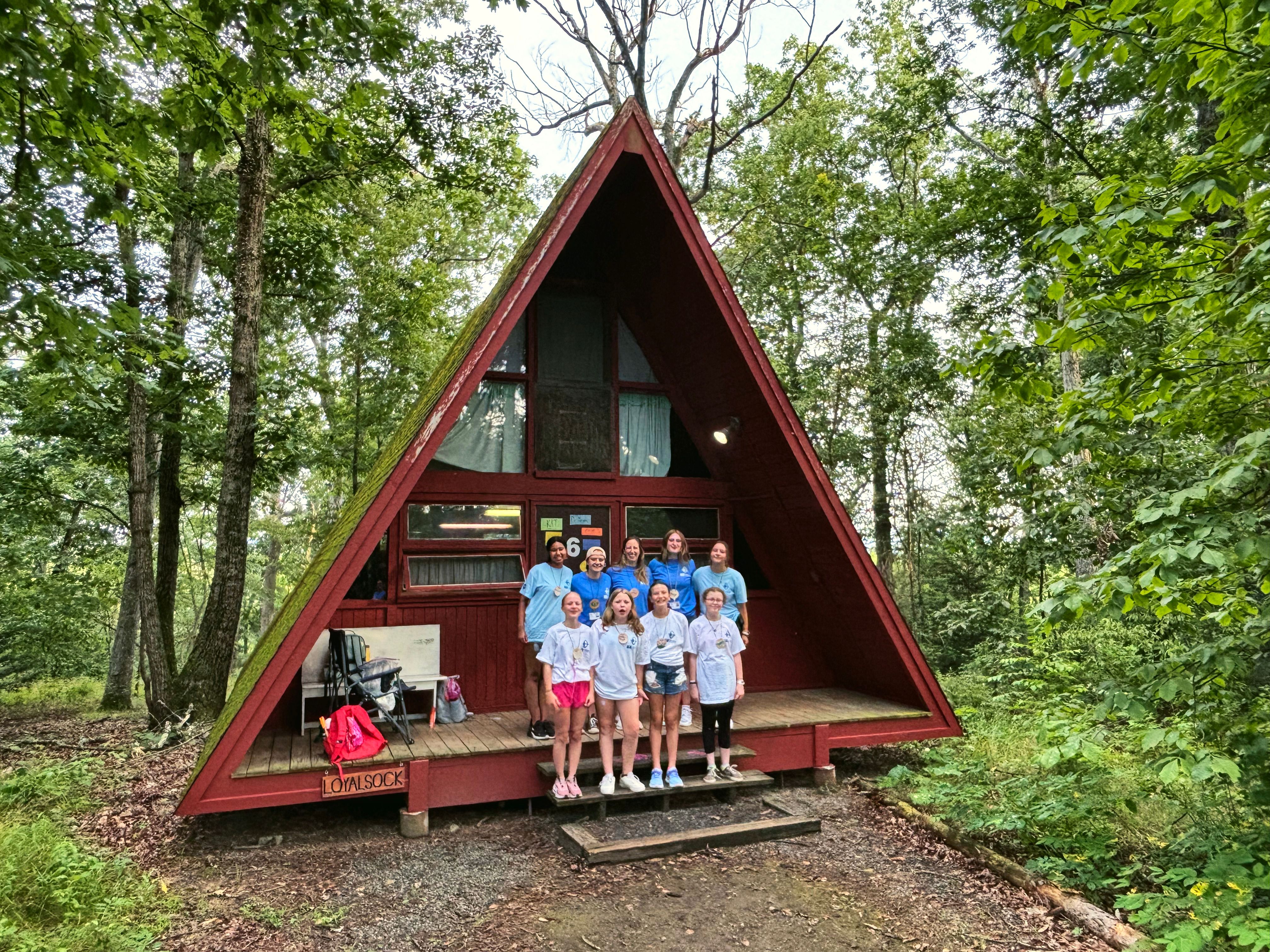 Campers stand together on the porch of a red A-frame cabin in the woods