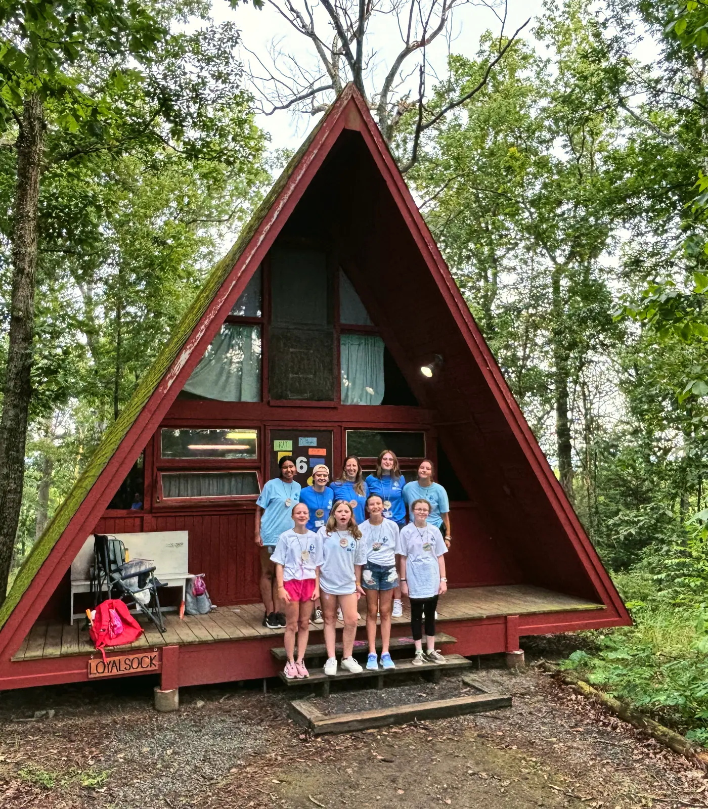 Campers stand together on the porch of a red A-frame cabin in the woods