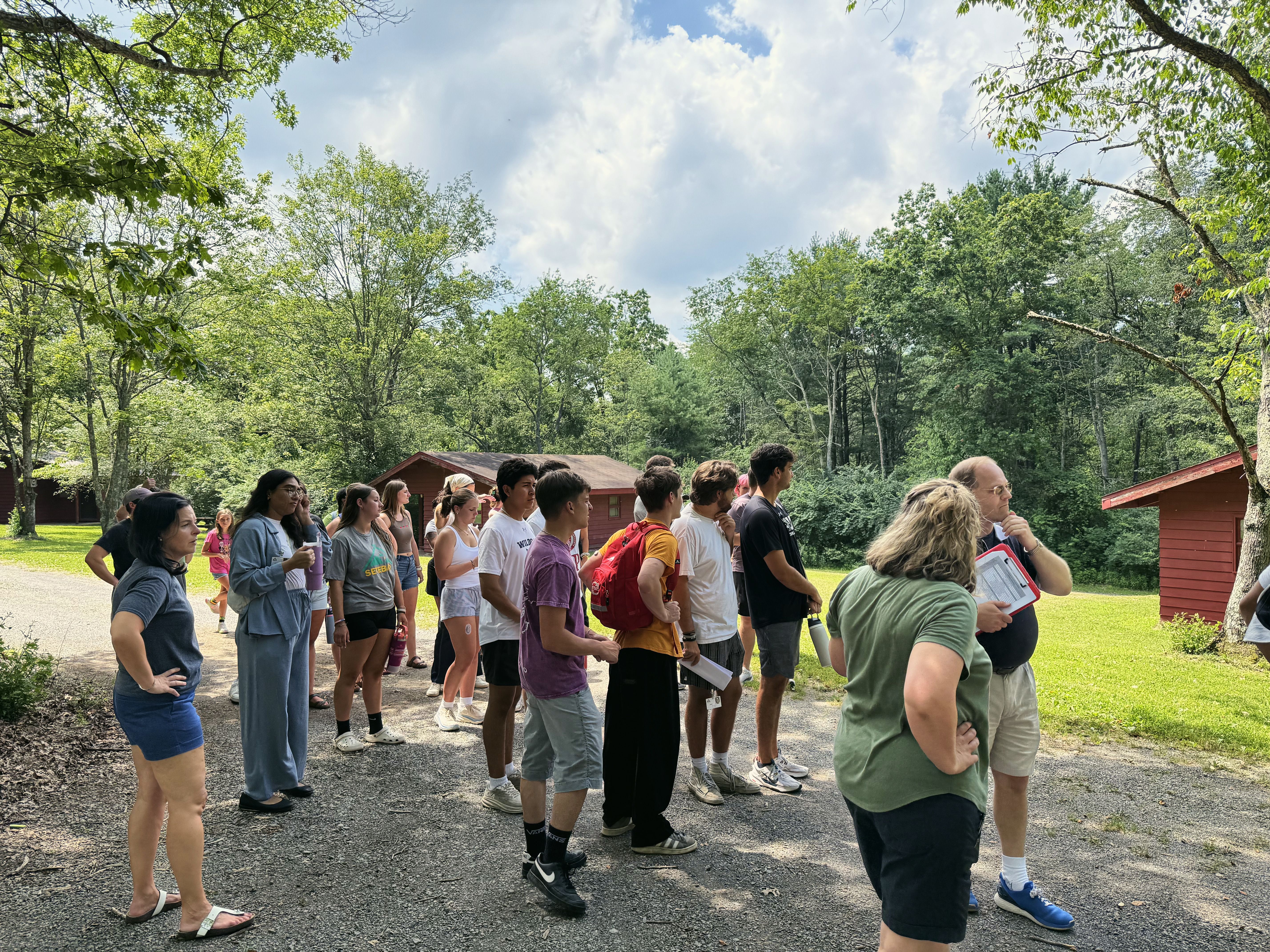 Teens and adults on a gravel path between cabins listen to a man giving instructions.