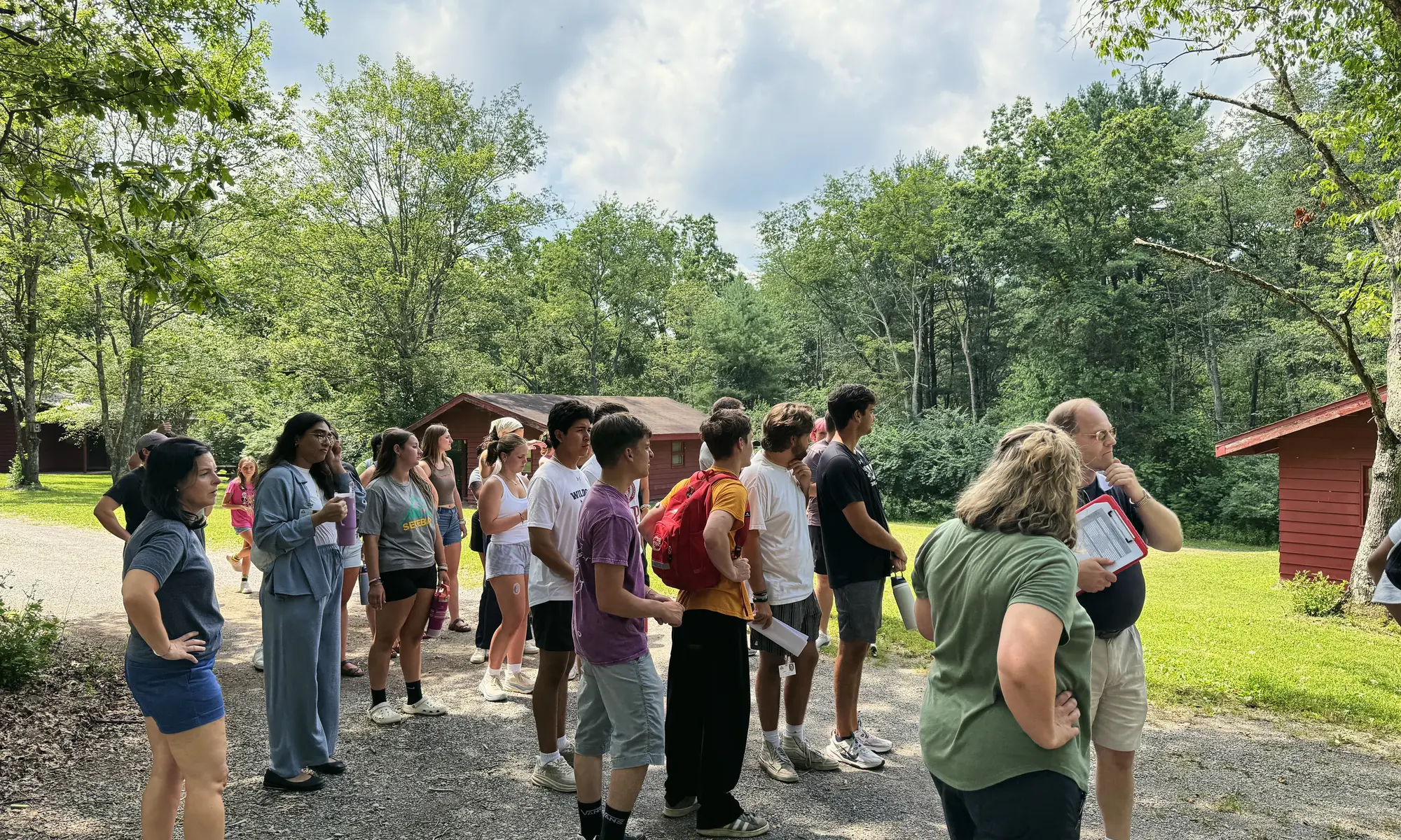 Teens and adults on a gravel path between cabins listen to a man giving instructions.
