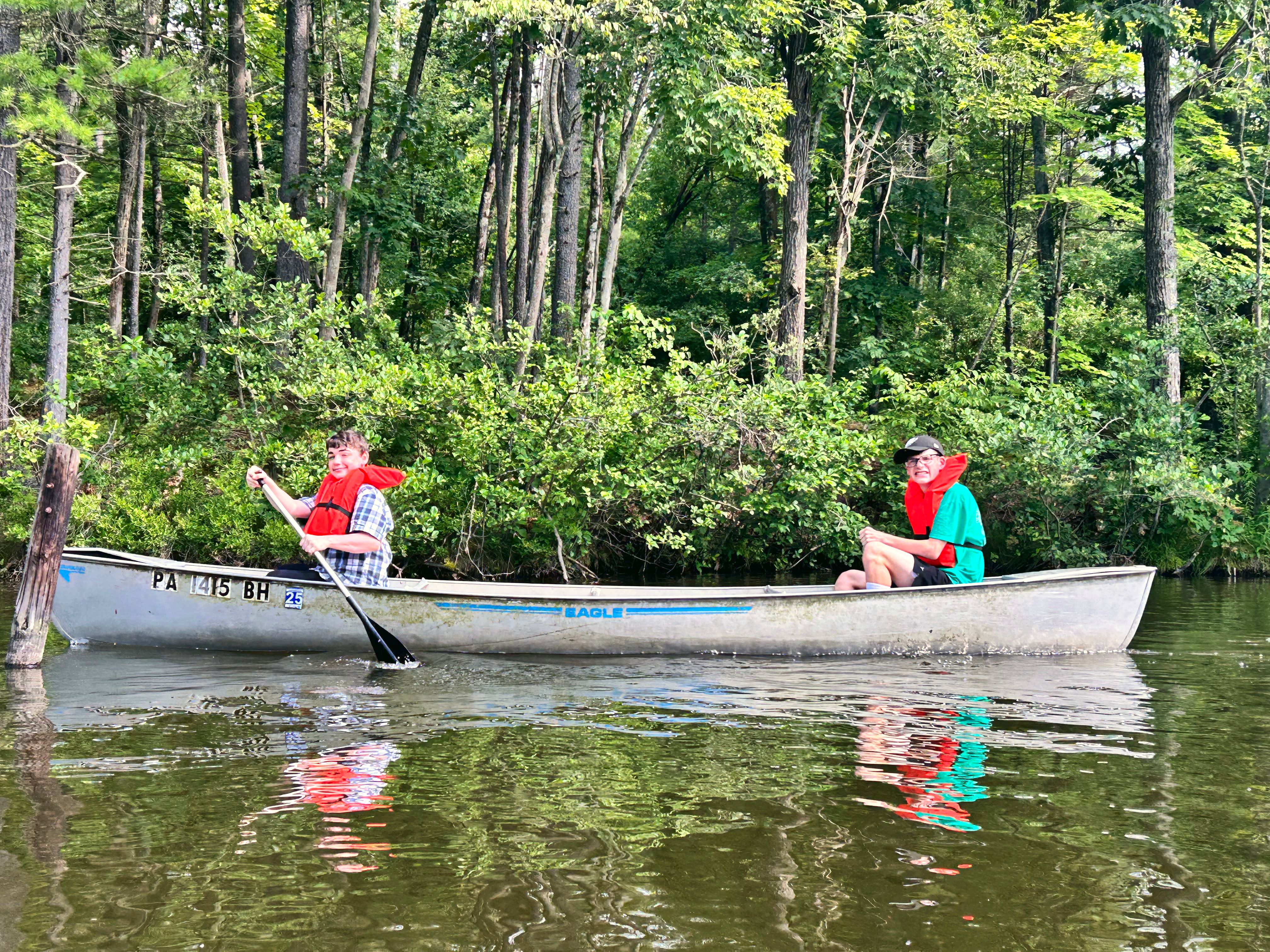 Two campers in life vests paddle a canoe on a calm river