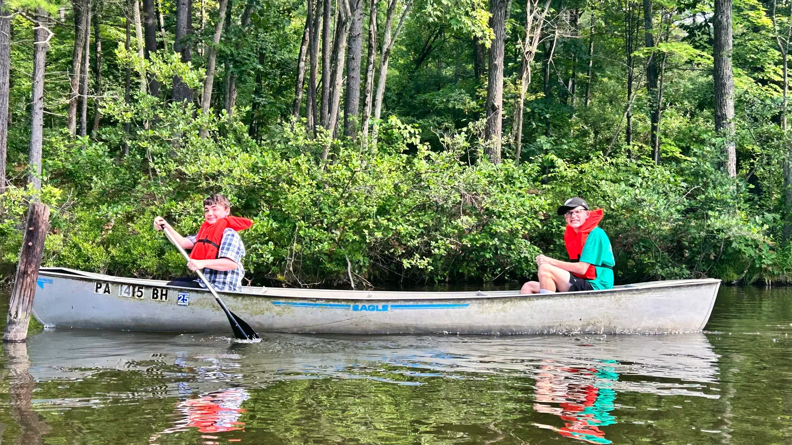 Two campers in life vests paddle a canoe on a calm river