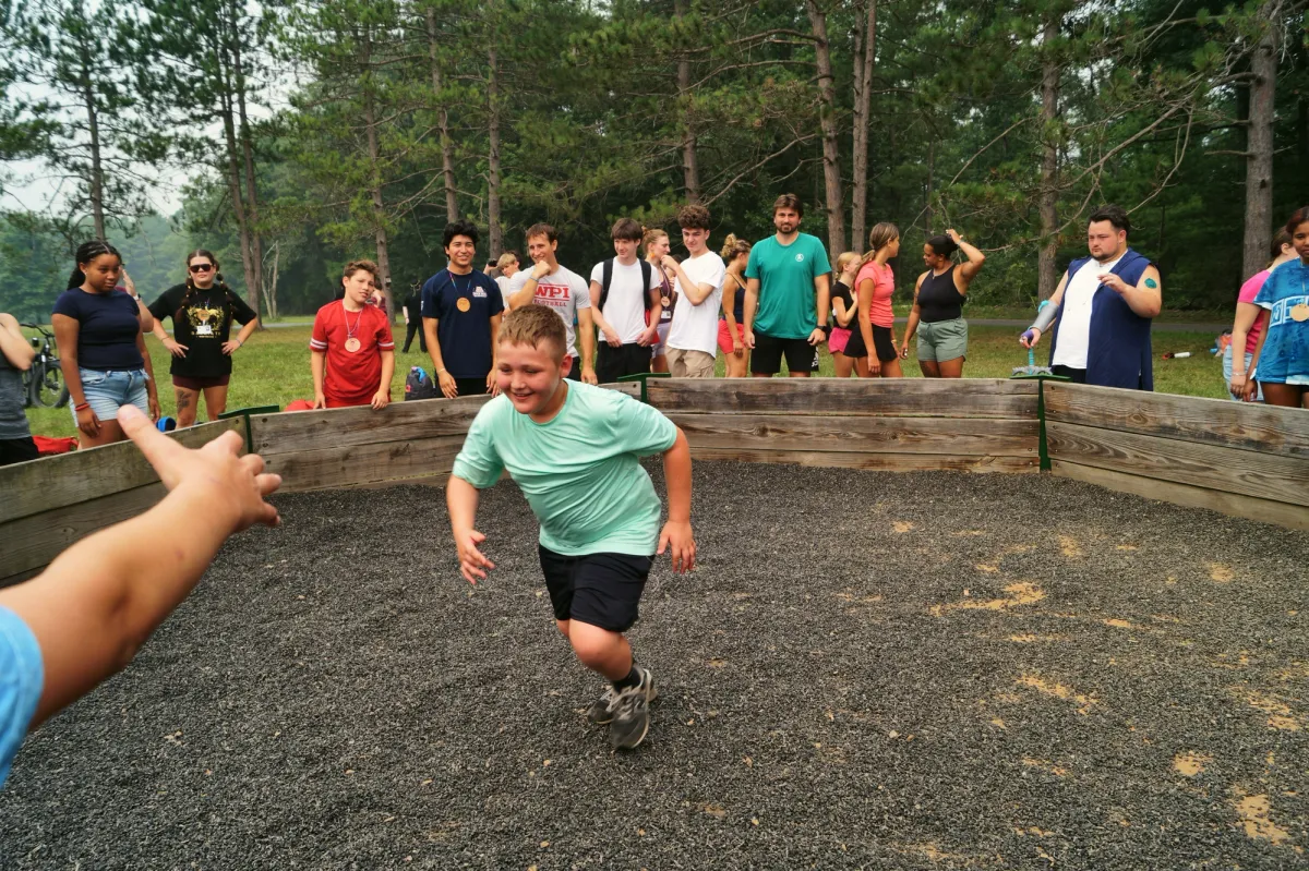 A smiling young camper runs across a gravel game pit while teens watch from the wooden wall