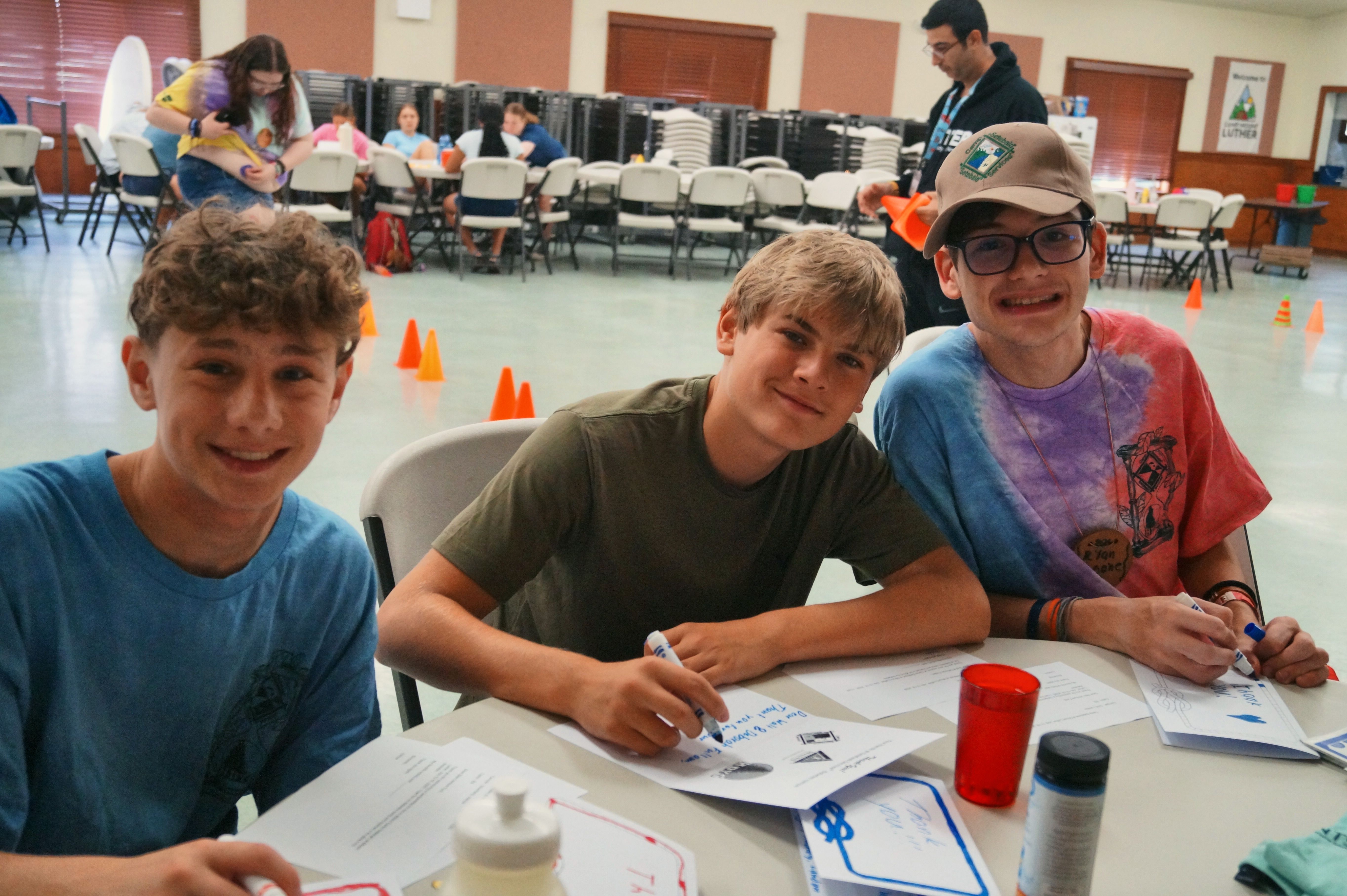 Three smiling boys sit together at a table in the camp dining hall.