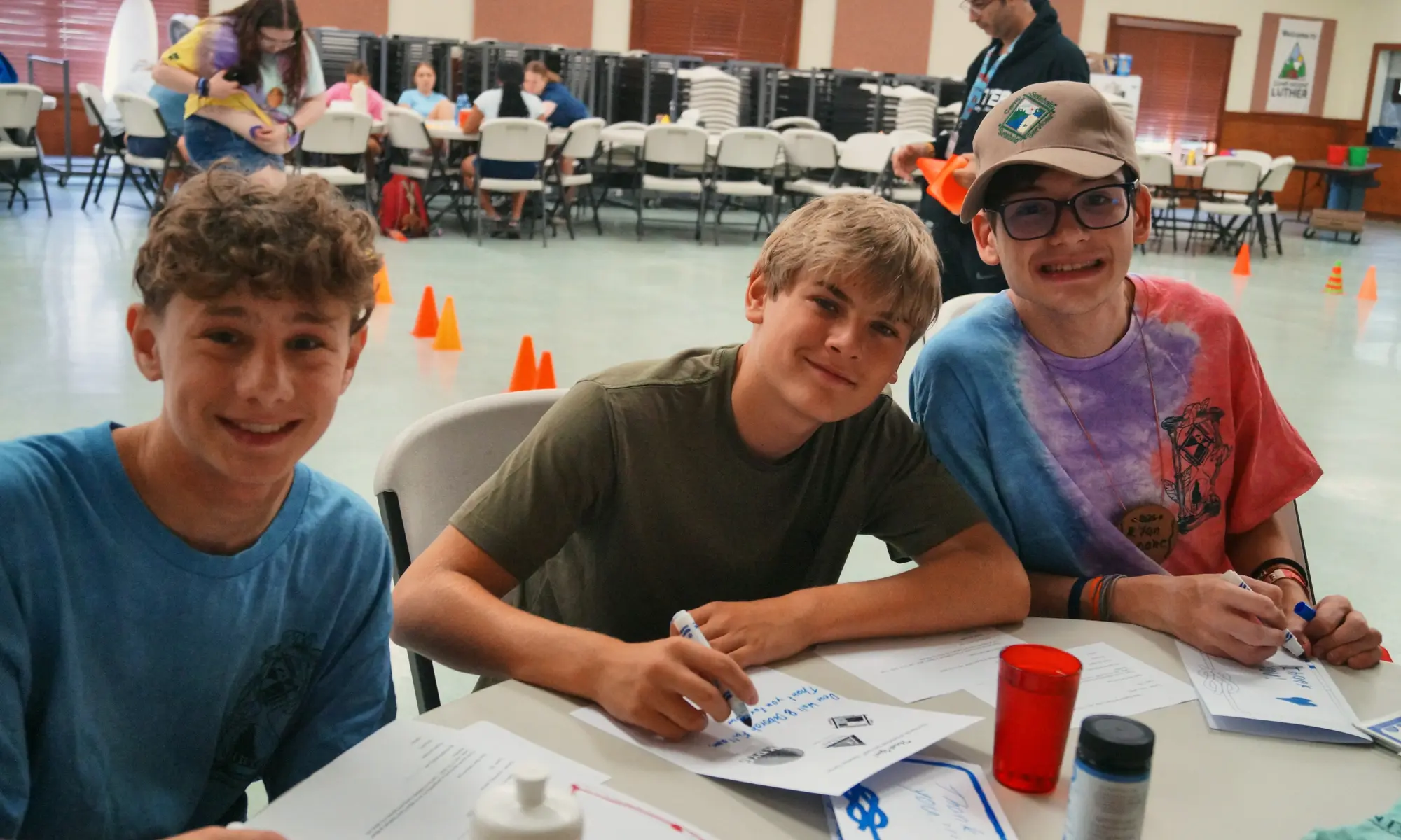 Three smiling boys sit together at a table in the camp dining hall.