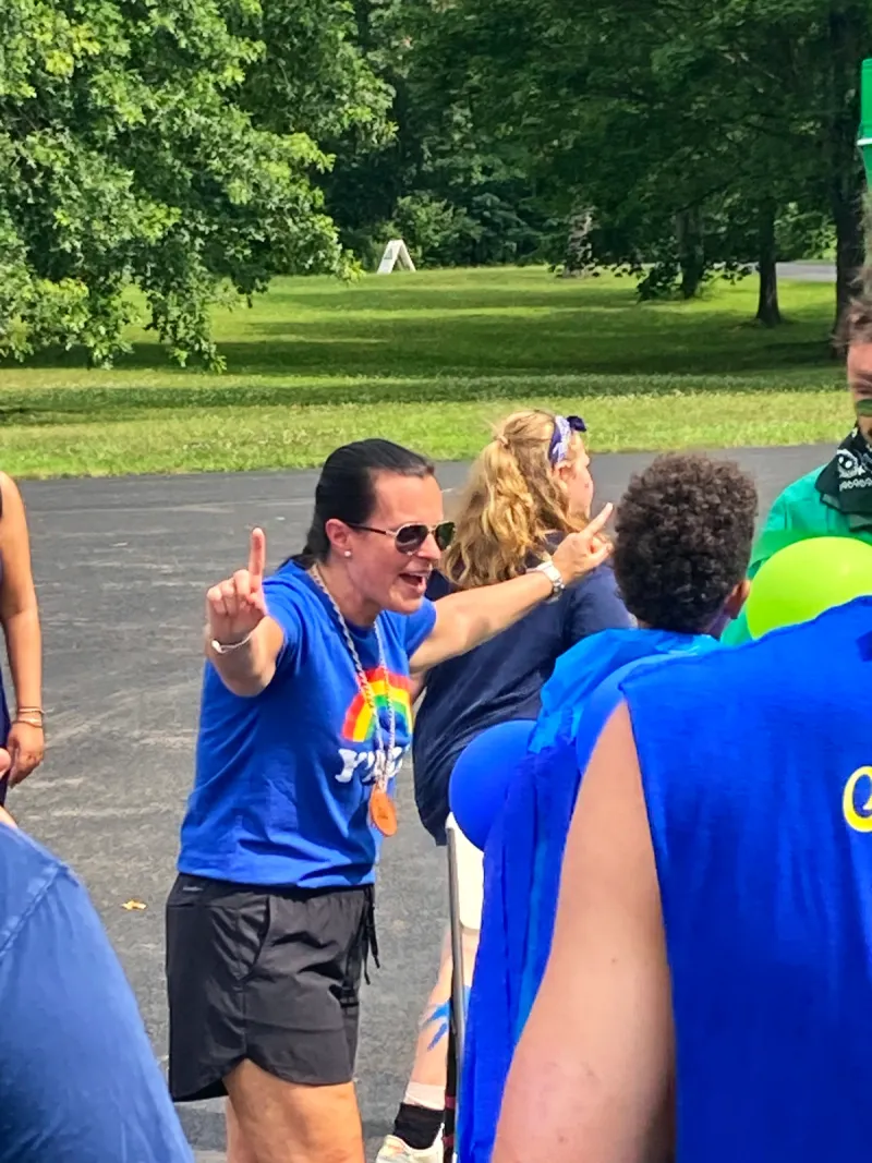A staff member in sunglasses and a rainbow shirt points upward as she talks to a group of campers.