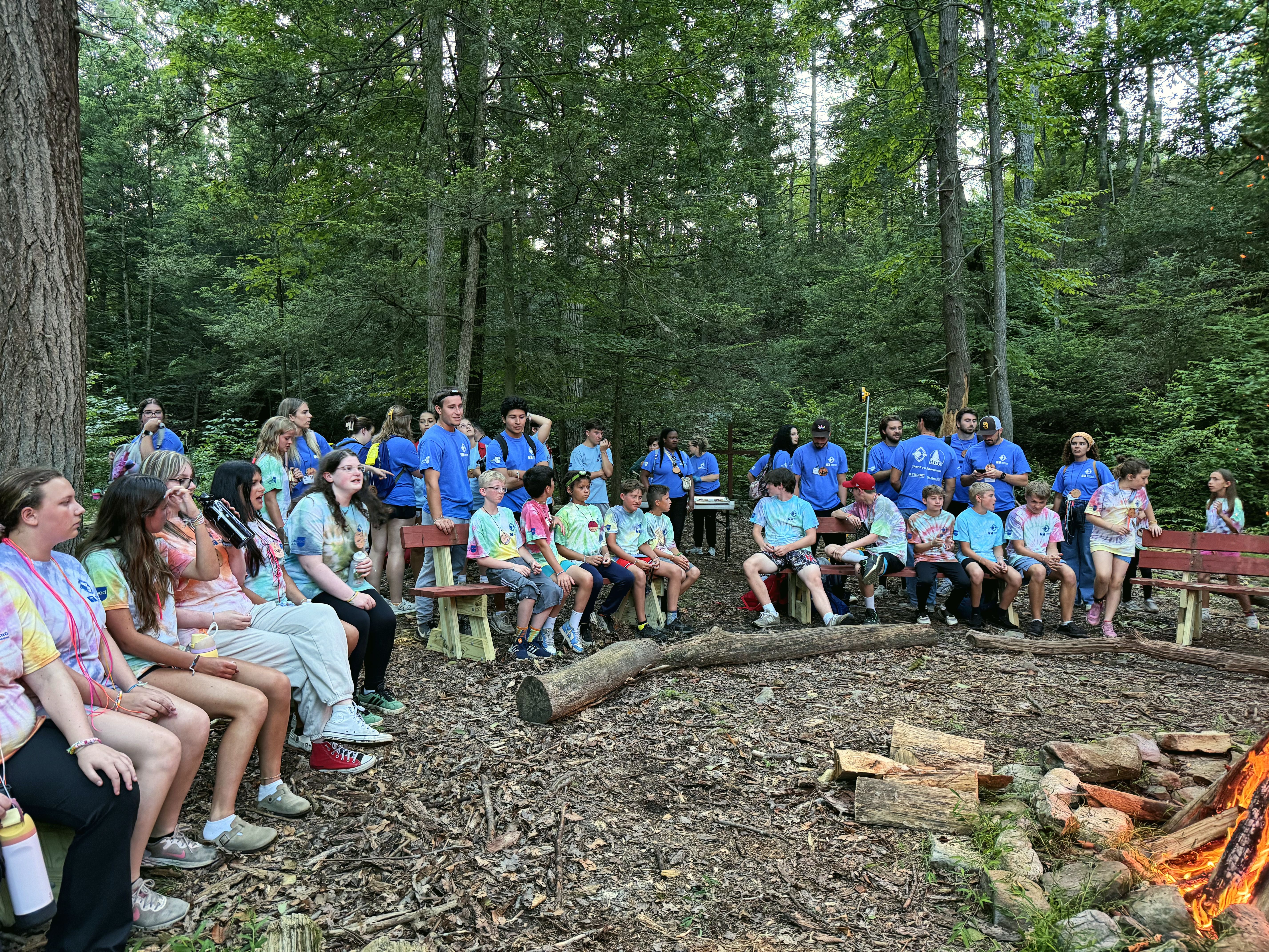 Campers and counselors sit together around a campfire circle in the woods