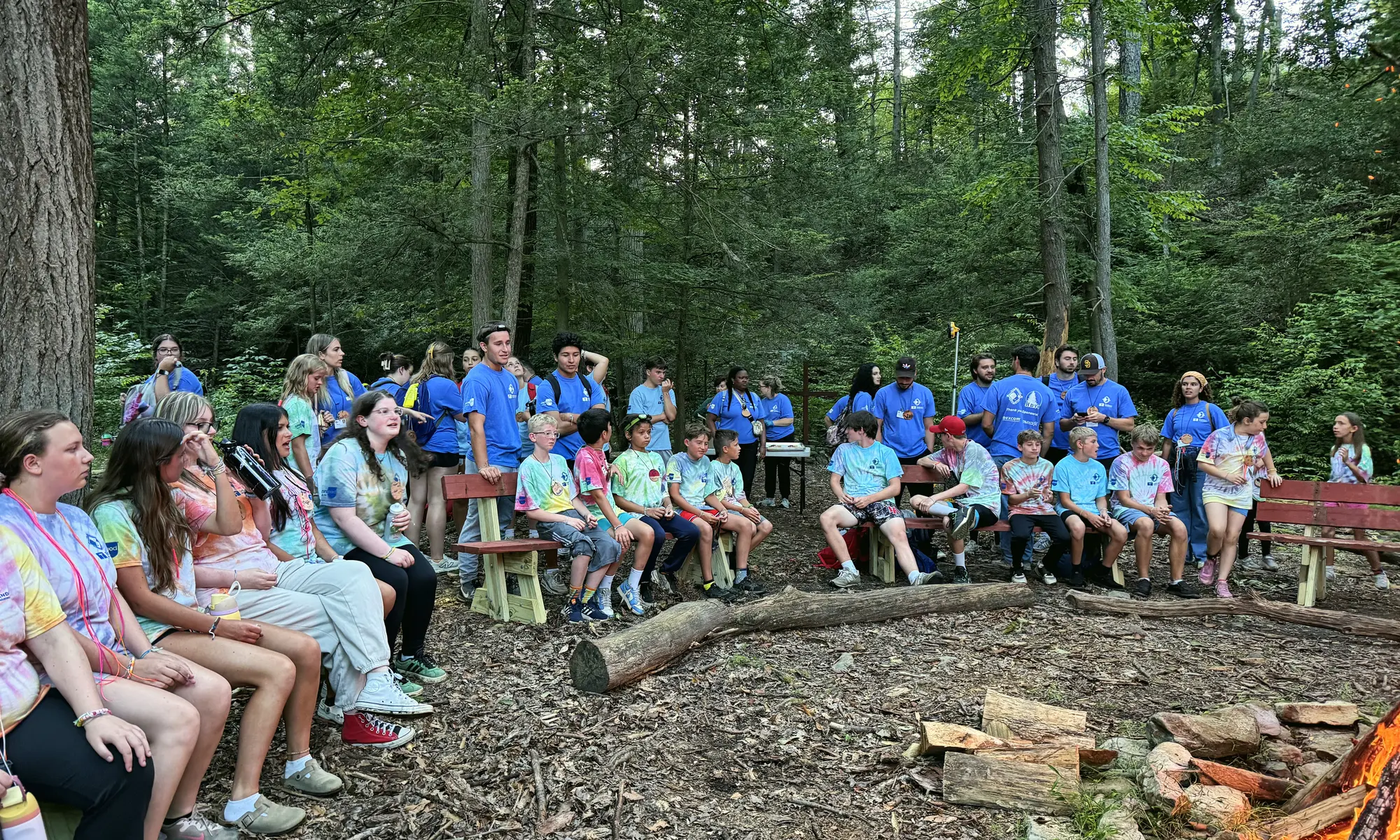 Campers and counselors sit together around a campfire circle in the woods