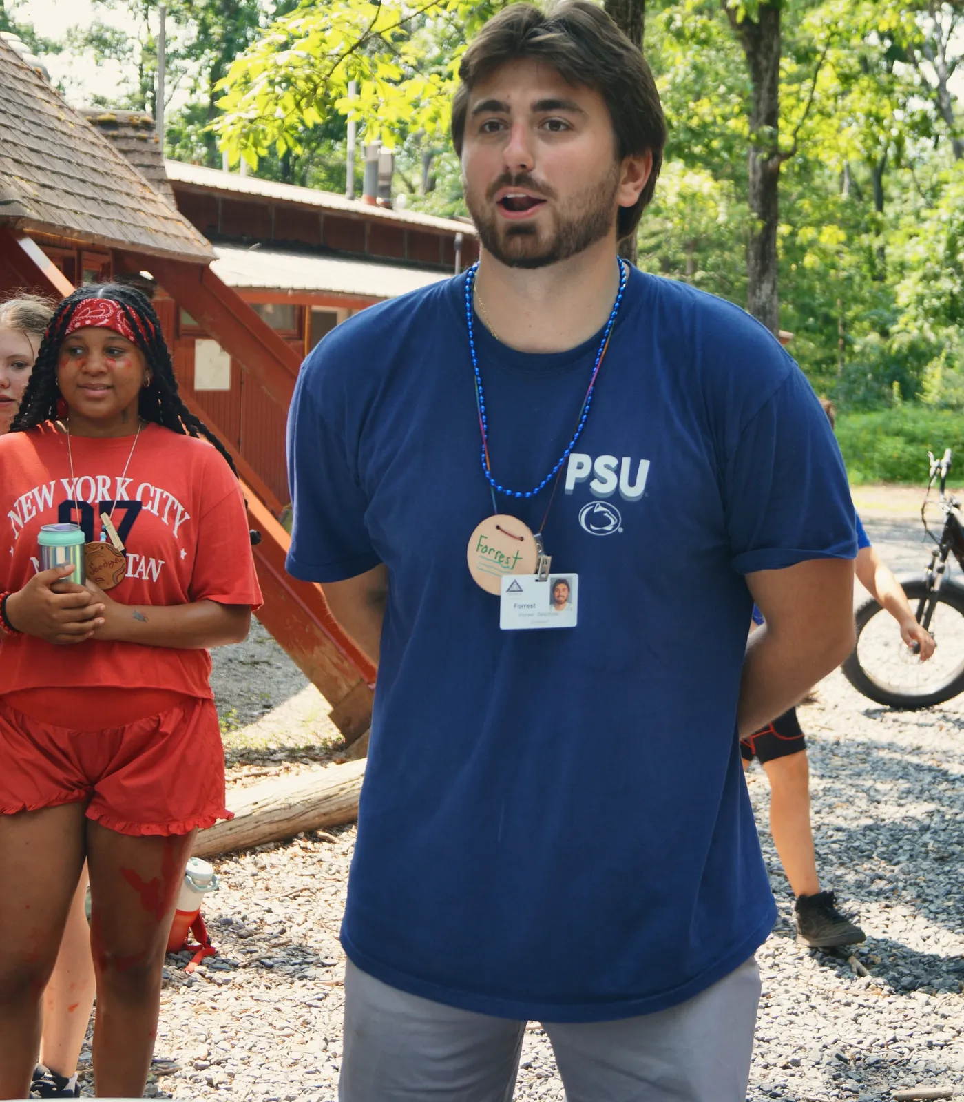 A young male counselor with a name badge talks to campers outside a cabin.