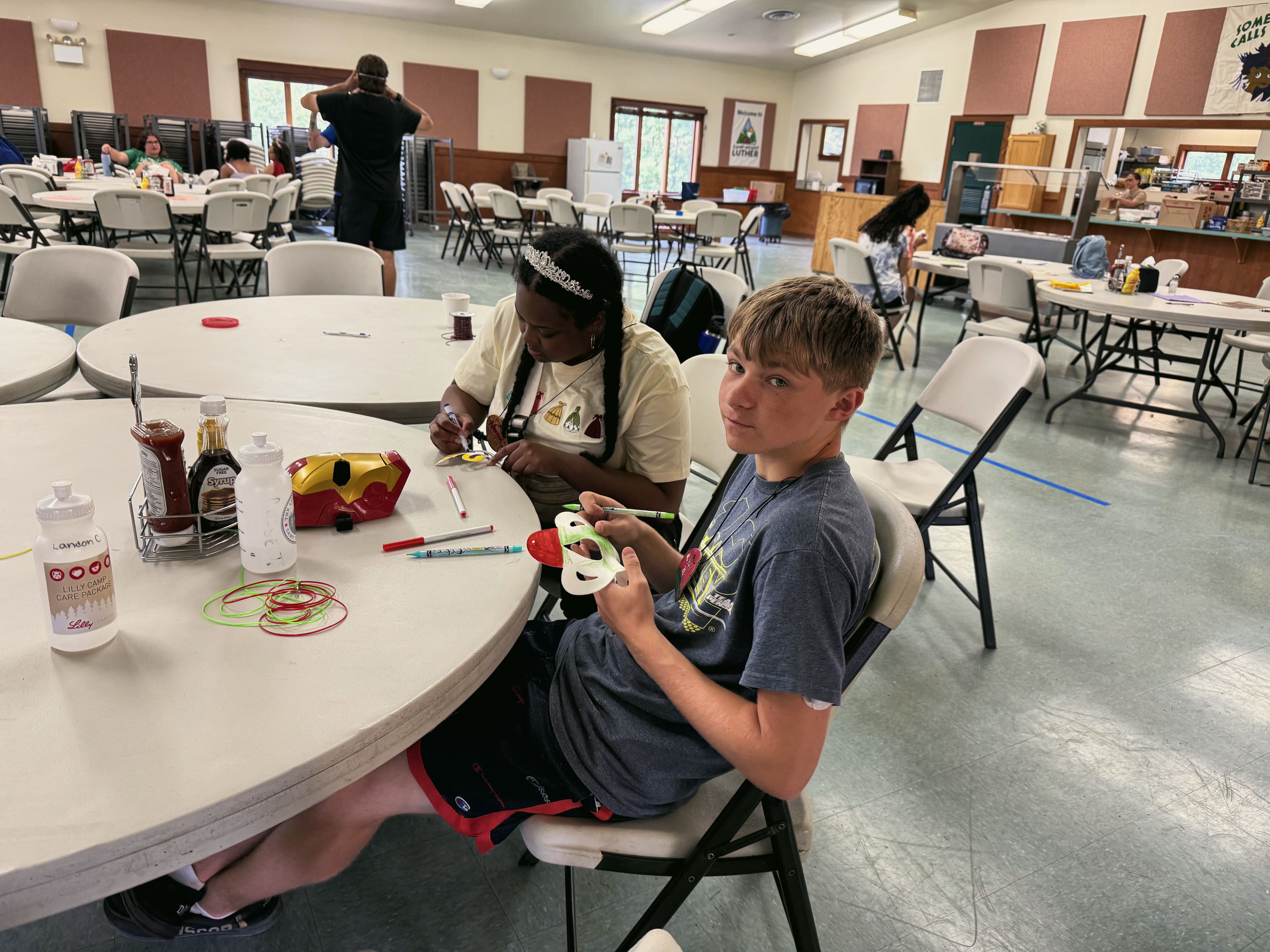 A boy shows his craft project at a round table in a busy hall, with a girl working beside him